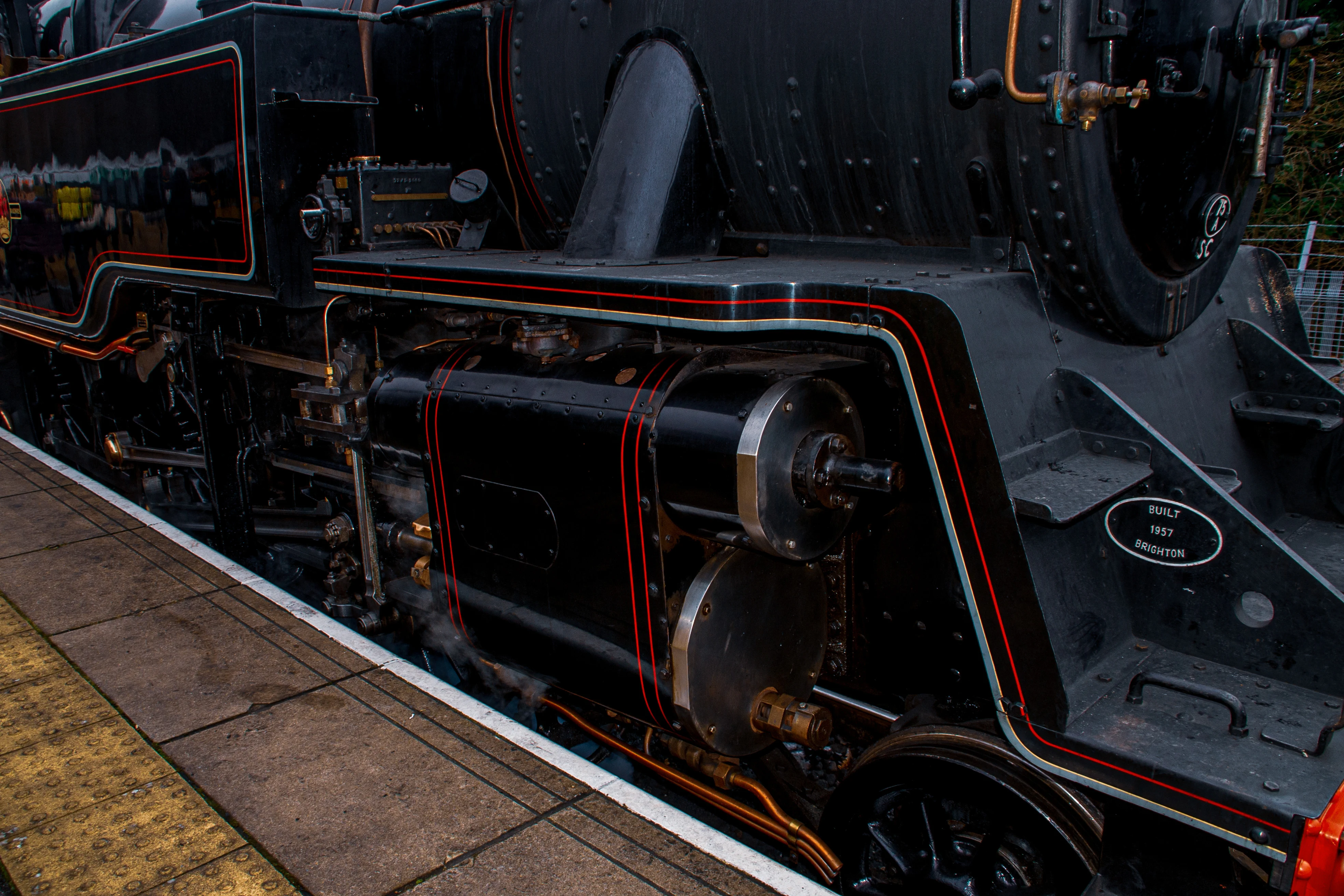 Close-up side view of a black steam locomotive, showing its detailed mechanics, pipes, and a plaque indicating it was built in 1957 in Brighton.