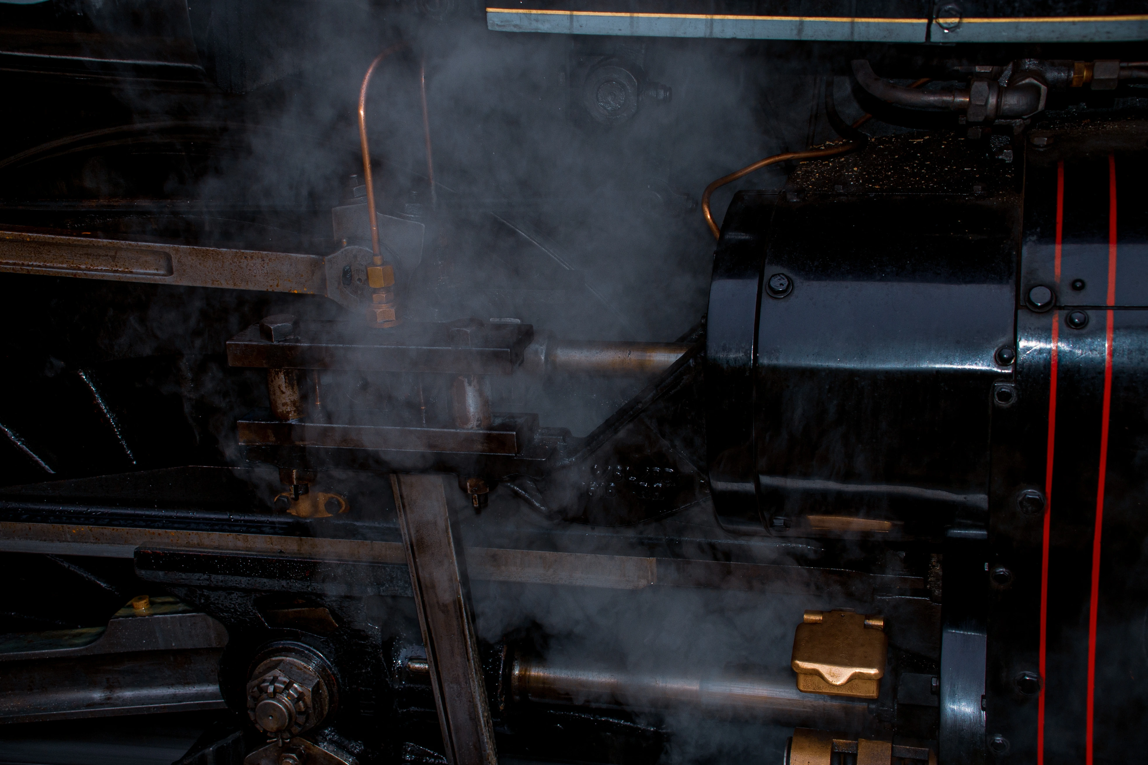 Close-up view of the complex mechanical parts of a steam train, with steam rising around dark metal levers, pipes, and a polished black engine casing with red stripe details.