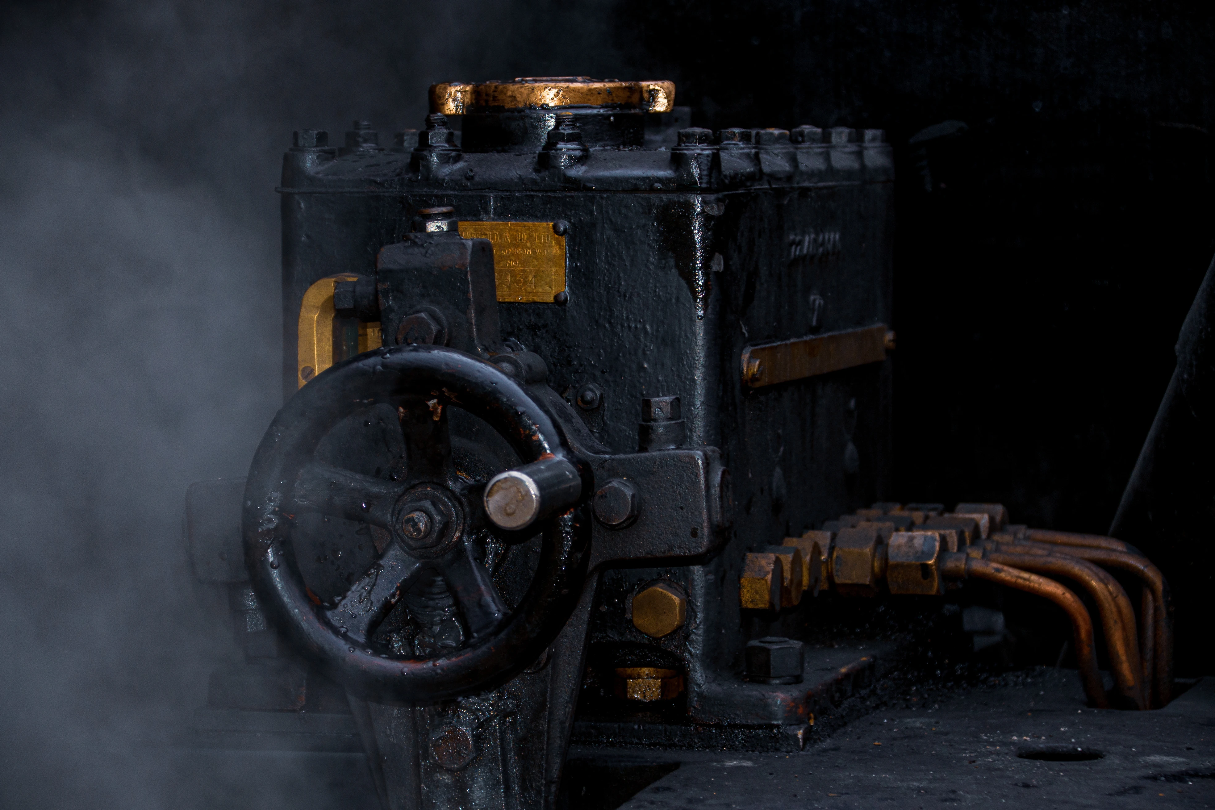 Detailed shot of a dark, industrial machine, possibly part of a steam engine, with a black metal body, a large black handwheel, and brass fittings. Wisps of steam drift across the left side of the frame, highlighting the machinery.