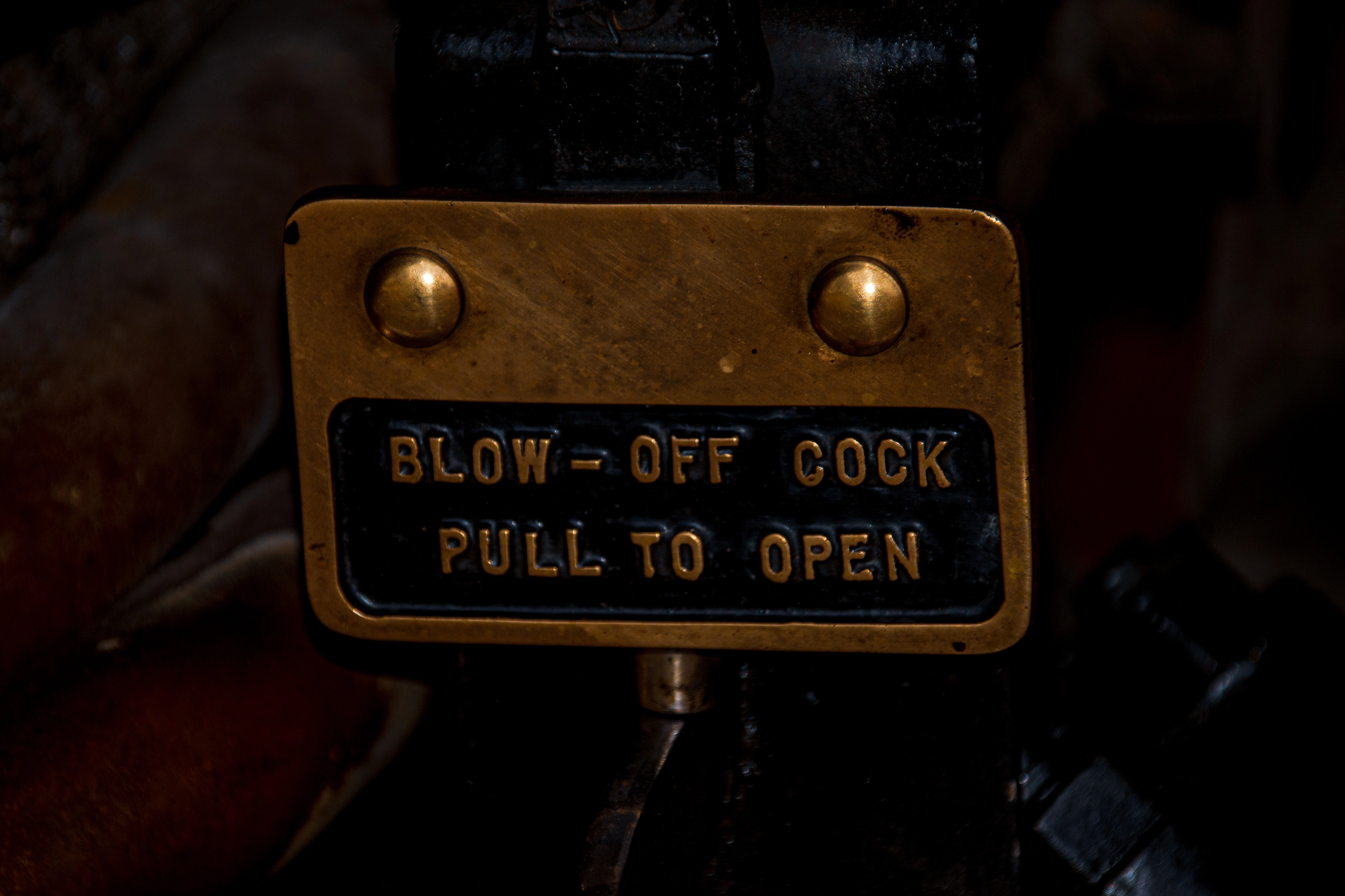 Close-up view of industrial machinery, possibly from a steam train, with wisps of steam obscuring parts of the dark metal gears, pipes, and bolts.