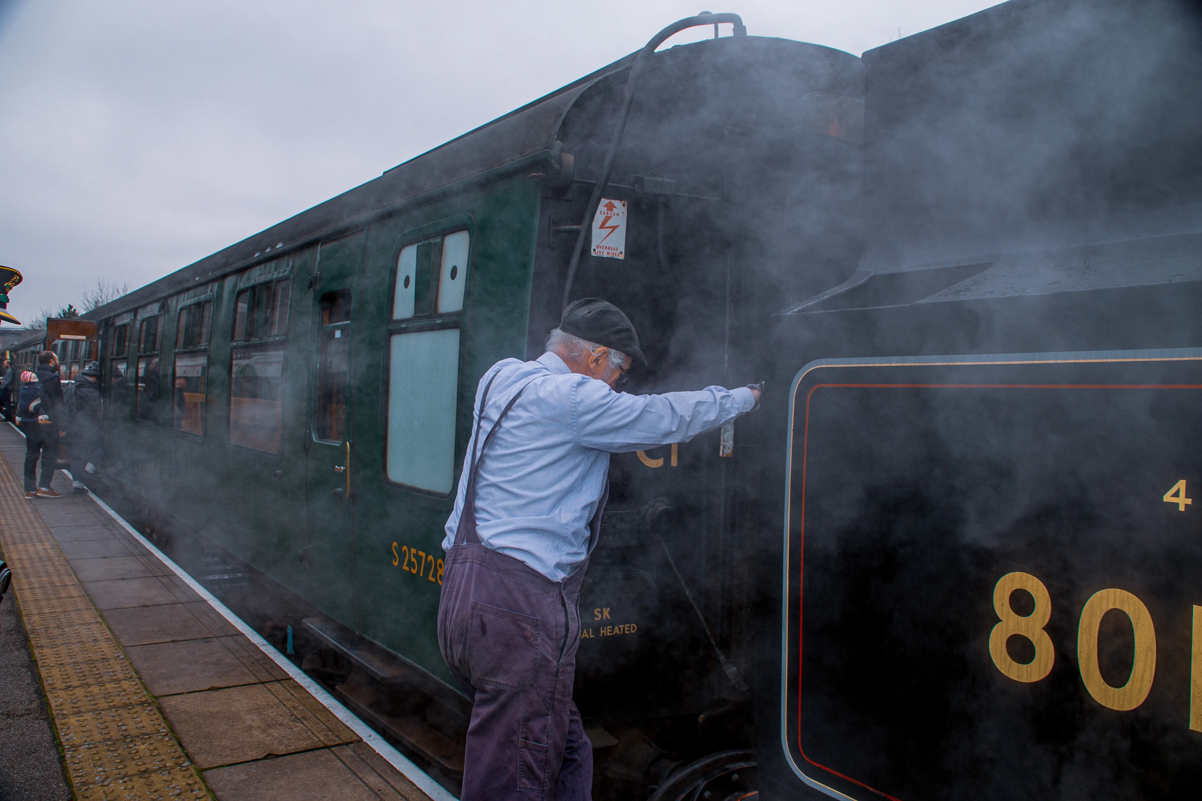 A train guard in overalls and a cap leans on a dark green railway carriage, surrounded by plumes of steam.