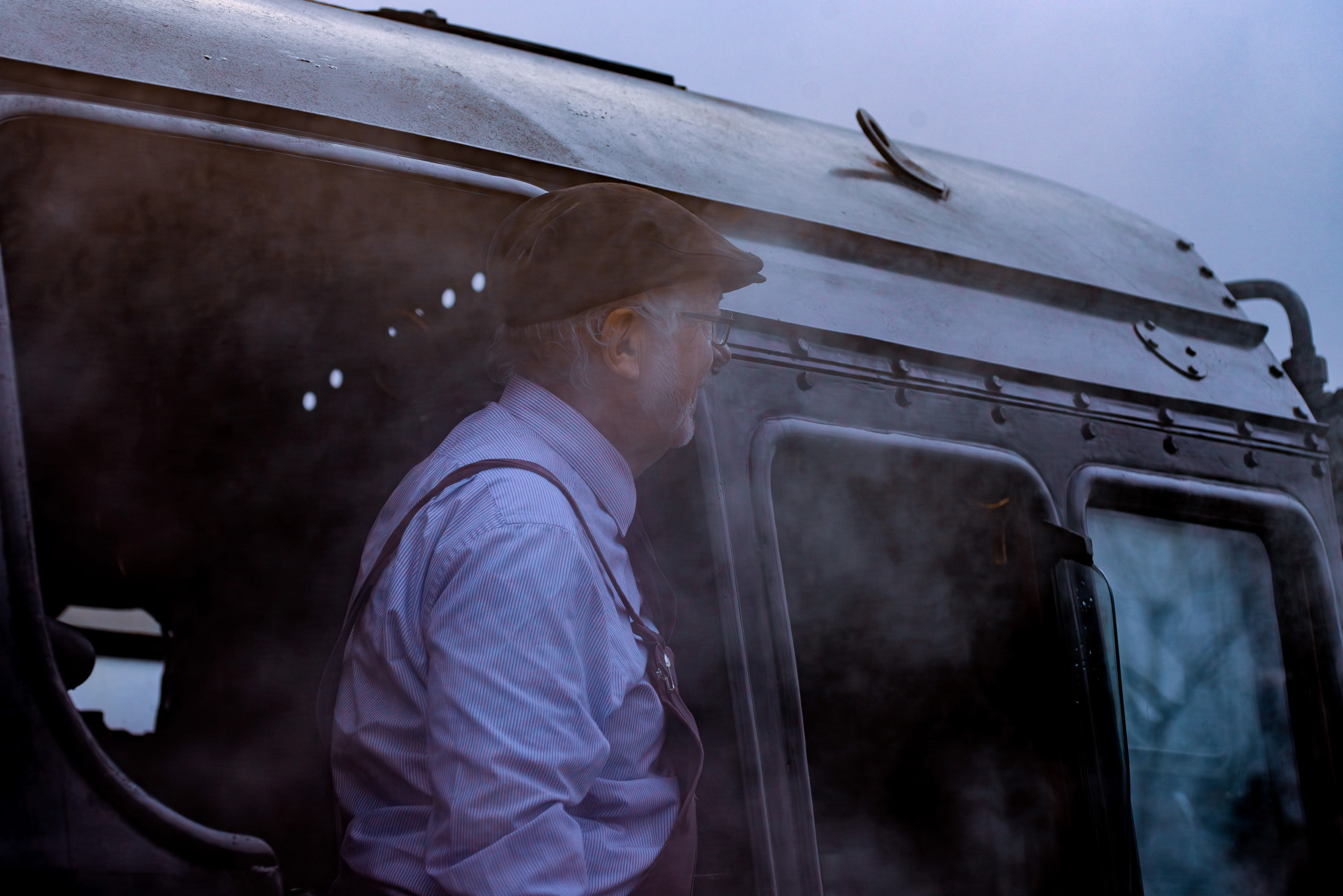 A man in a cap and braces looks out of a steam train's cab, with steam obscuring parts of the locomotive.