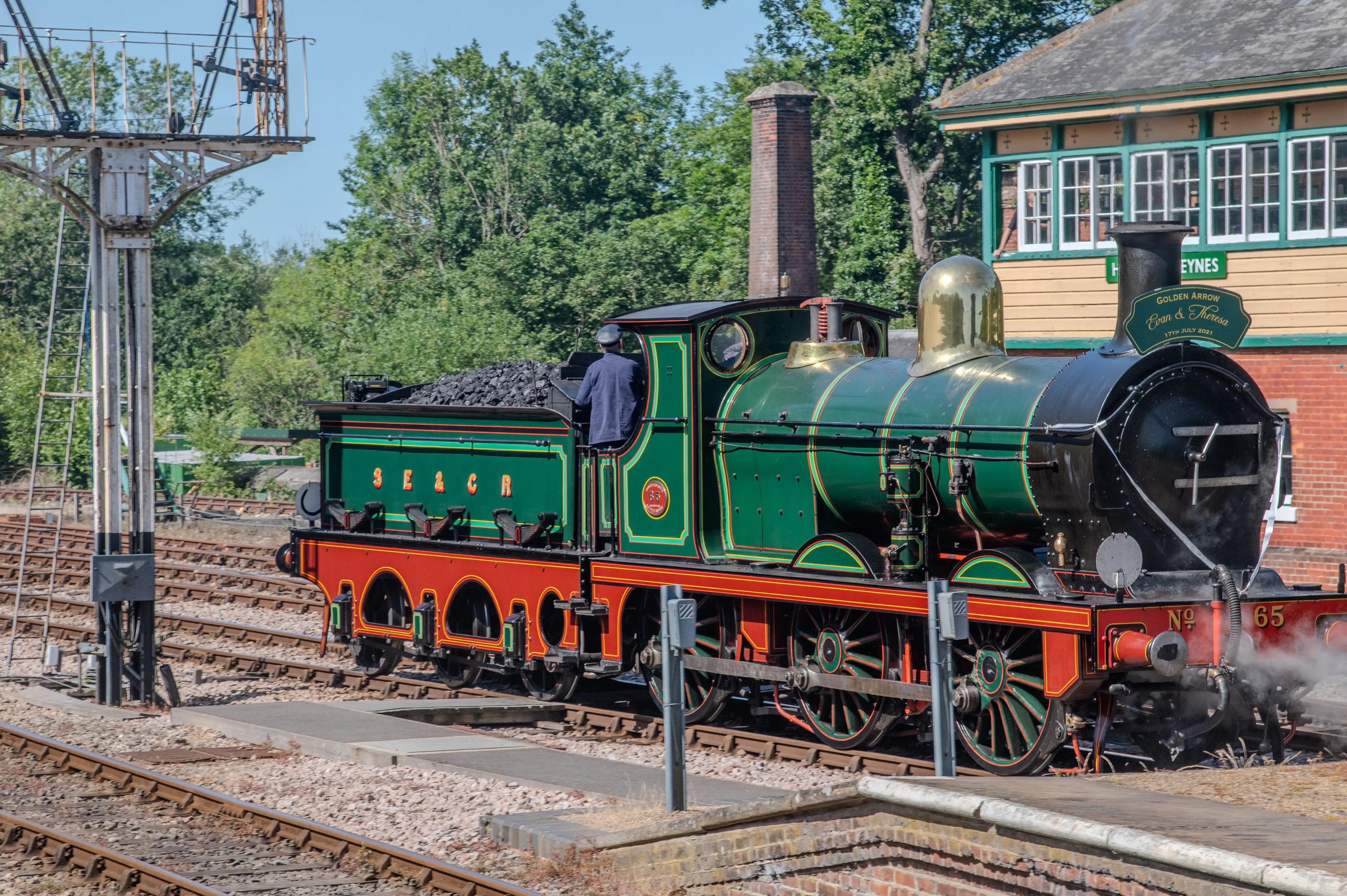 A beautifully restored vintage steam locomotive, No. 65, painted in green and red with gold accents, pulling a coal tender marked 'SE&CR'. The locomotive is in motion, emitting steam, and is operated by a person in the cabin. The scene is set at a railway station with multiple tracks, a signal post, and a station building labeled 'Horsted Keynes' in the background. The date '17th July 2021' is visible on a sign