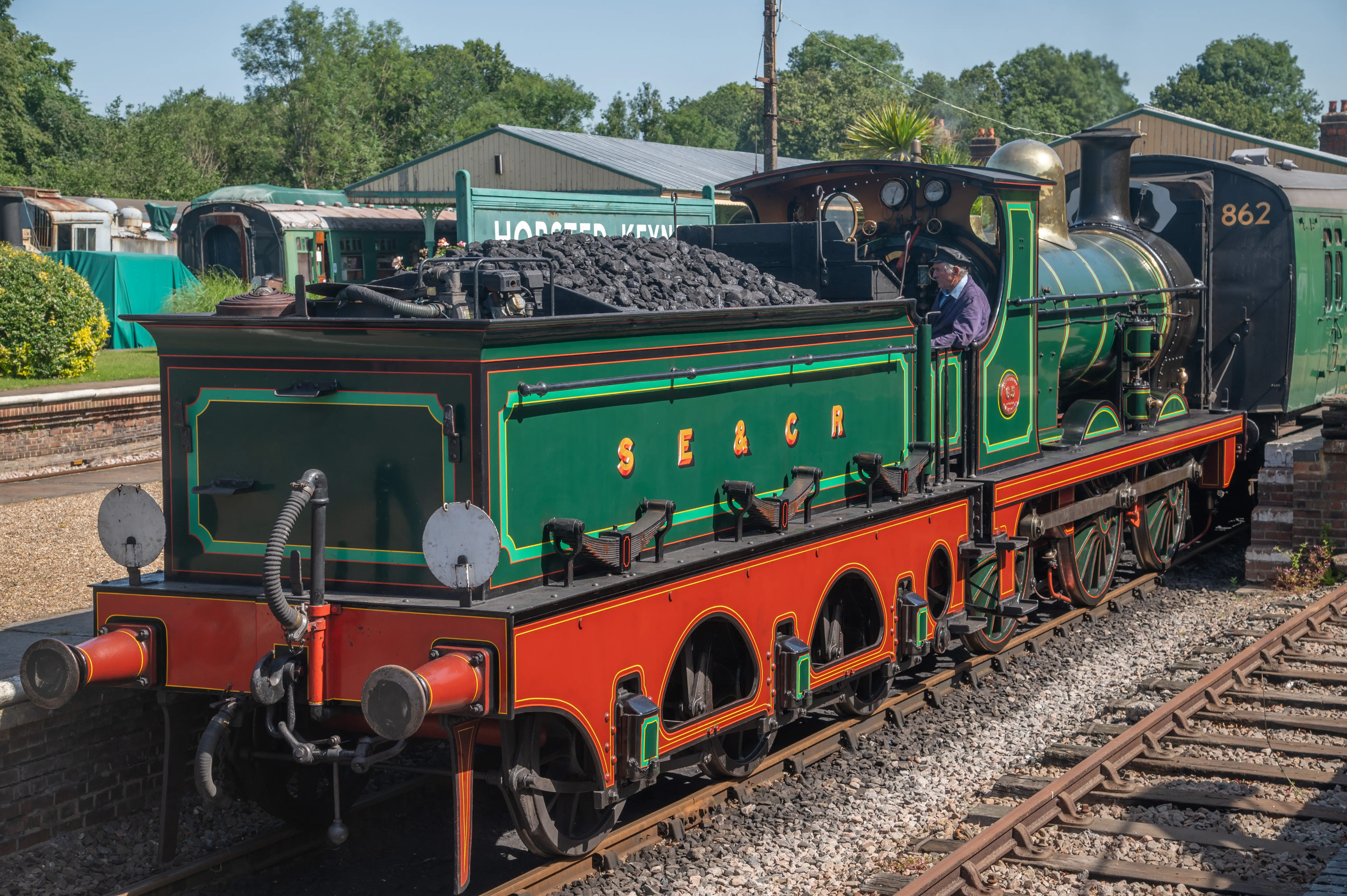 A green and red steam locomotive with S.E. & C.R. emblazoned on its side, number 862 on the tender, stands at a station platform. A driver in a cap is visible in the cab.