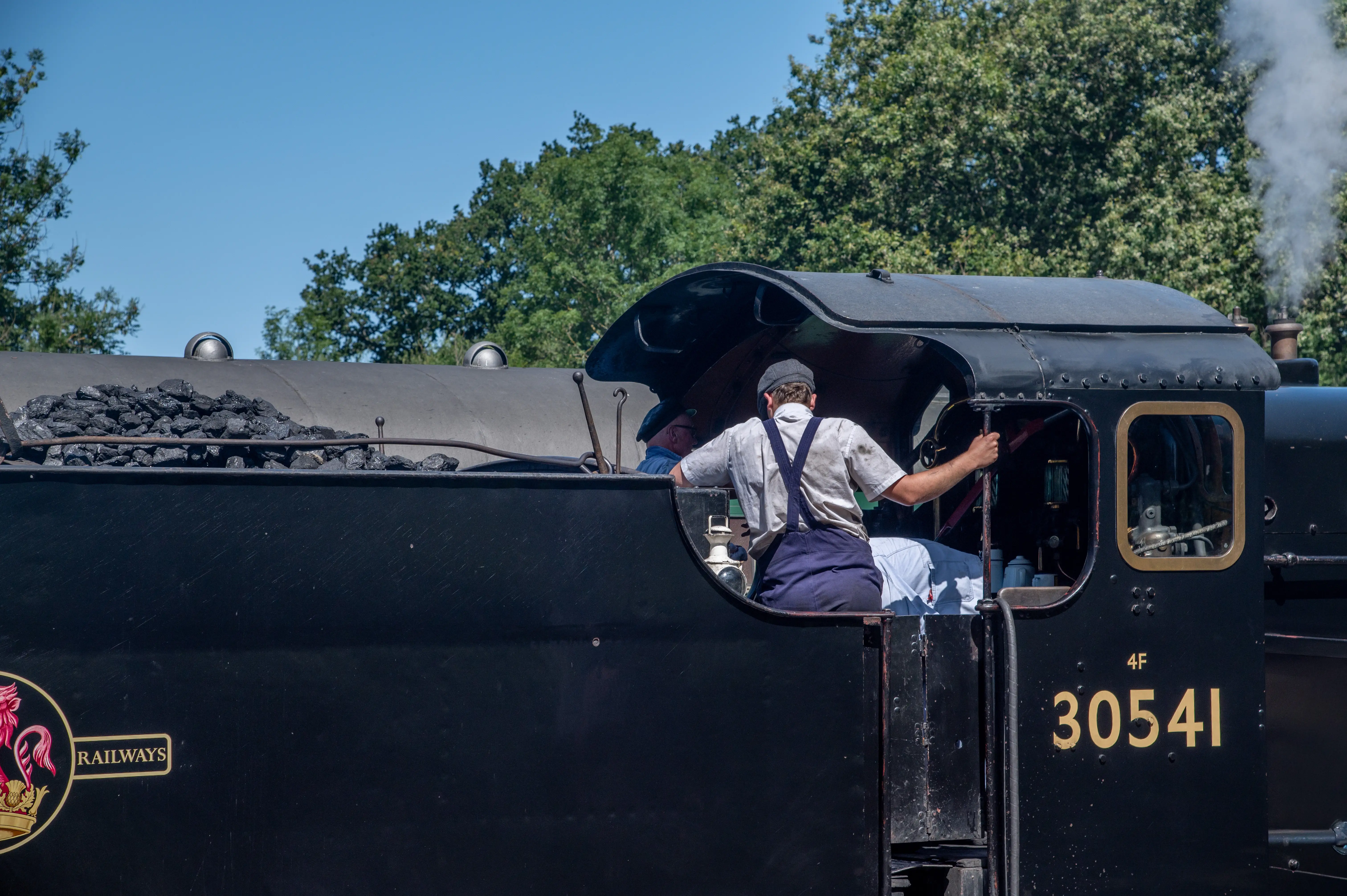 Two men in work overalls and caps are inside the cabin of a black steam train. One man, facing away from the camera, grips a lever. A pile of coal is visible in the foreground, and smoke rises from the train in the background.