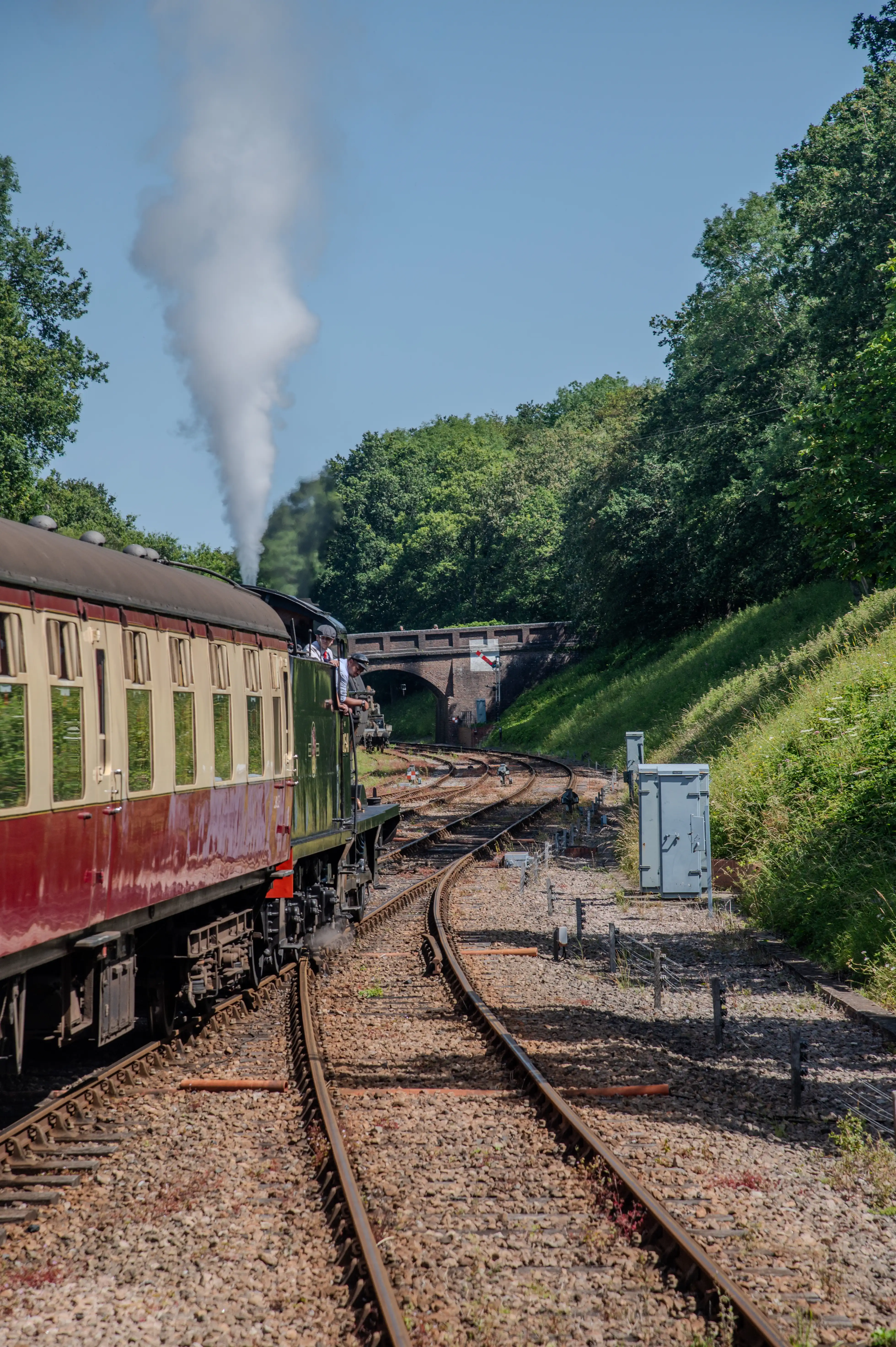 A side view of a red and cream vintage train with a dark green engine steaming heavily as it rounds a bend on a sunny day, with trees and a brick bridge in the background.