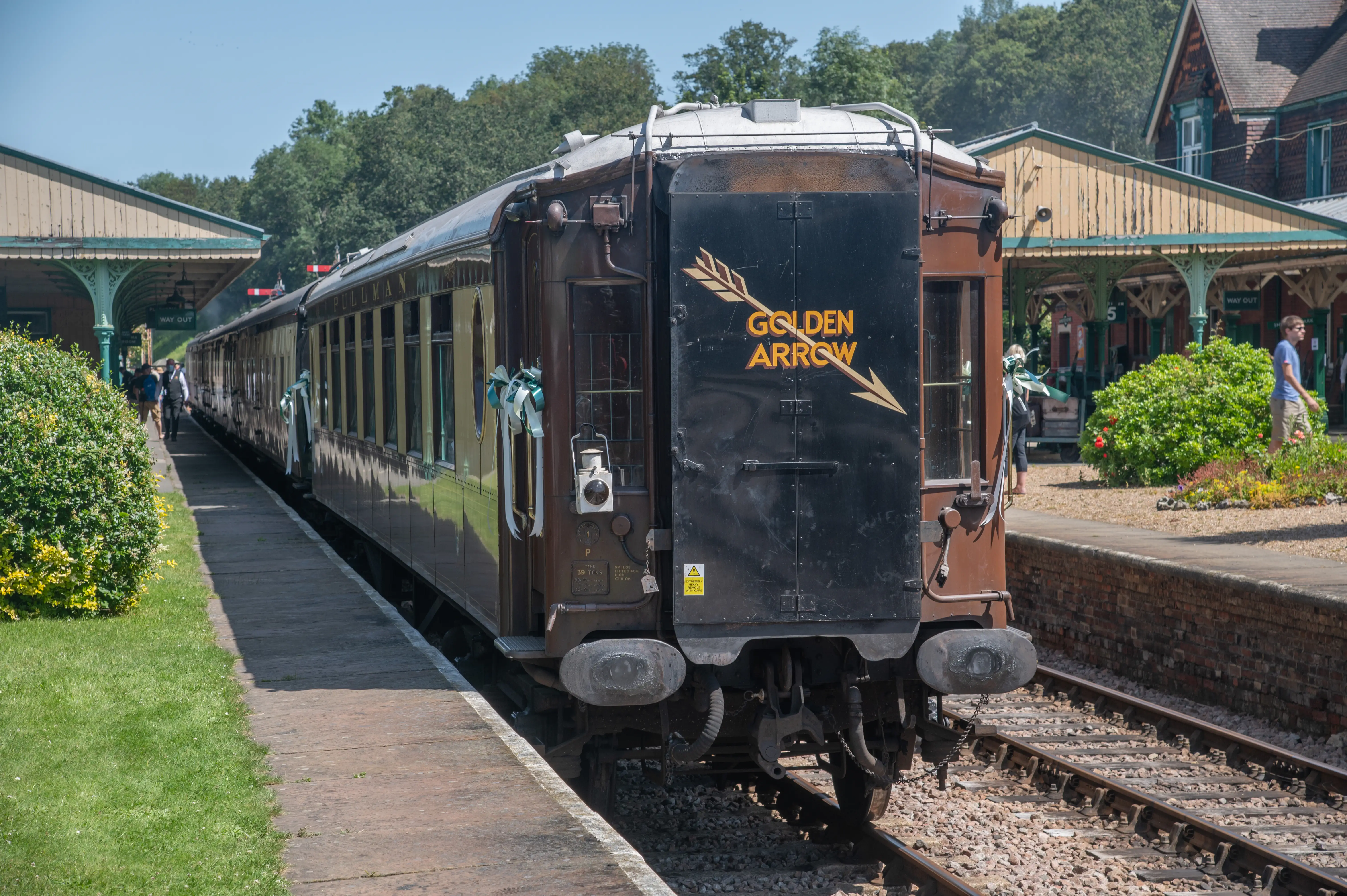 Rear view of a brown and gold Pullman train carriage bearing the 'Golden Arrow' logo, parked at a station platform with people and buildings in the background.