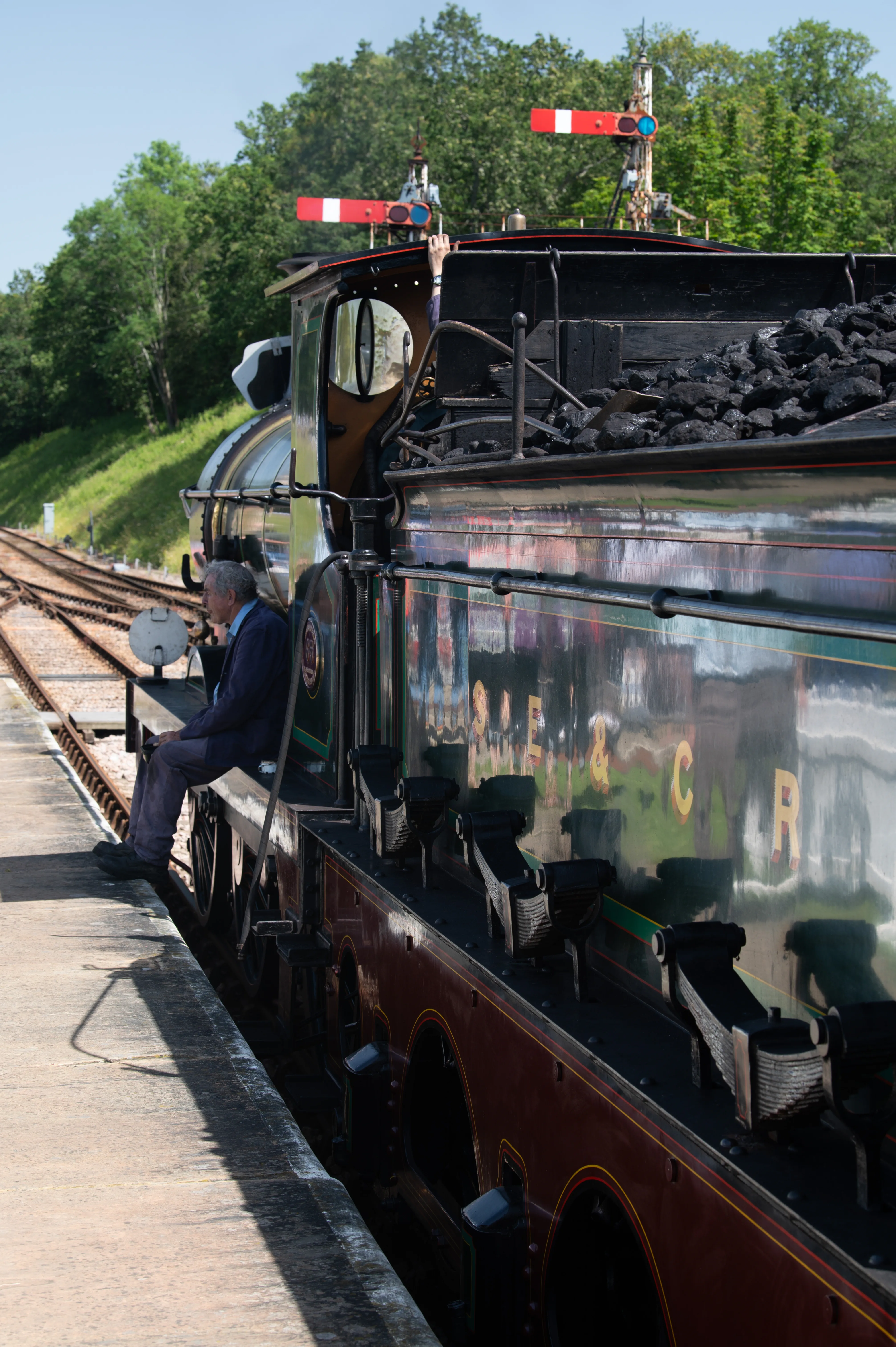 A side view of a vintage steam train with a man sitting on the edge of its footplate, waiting.
