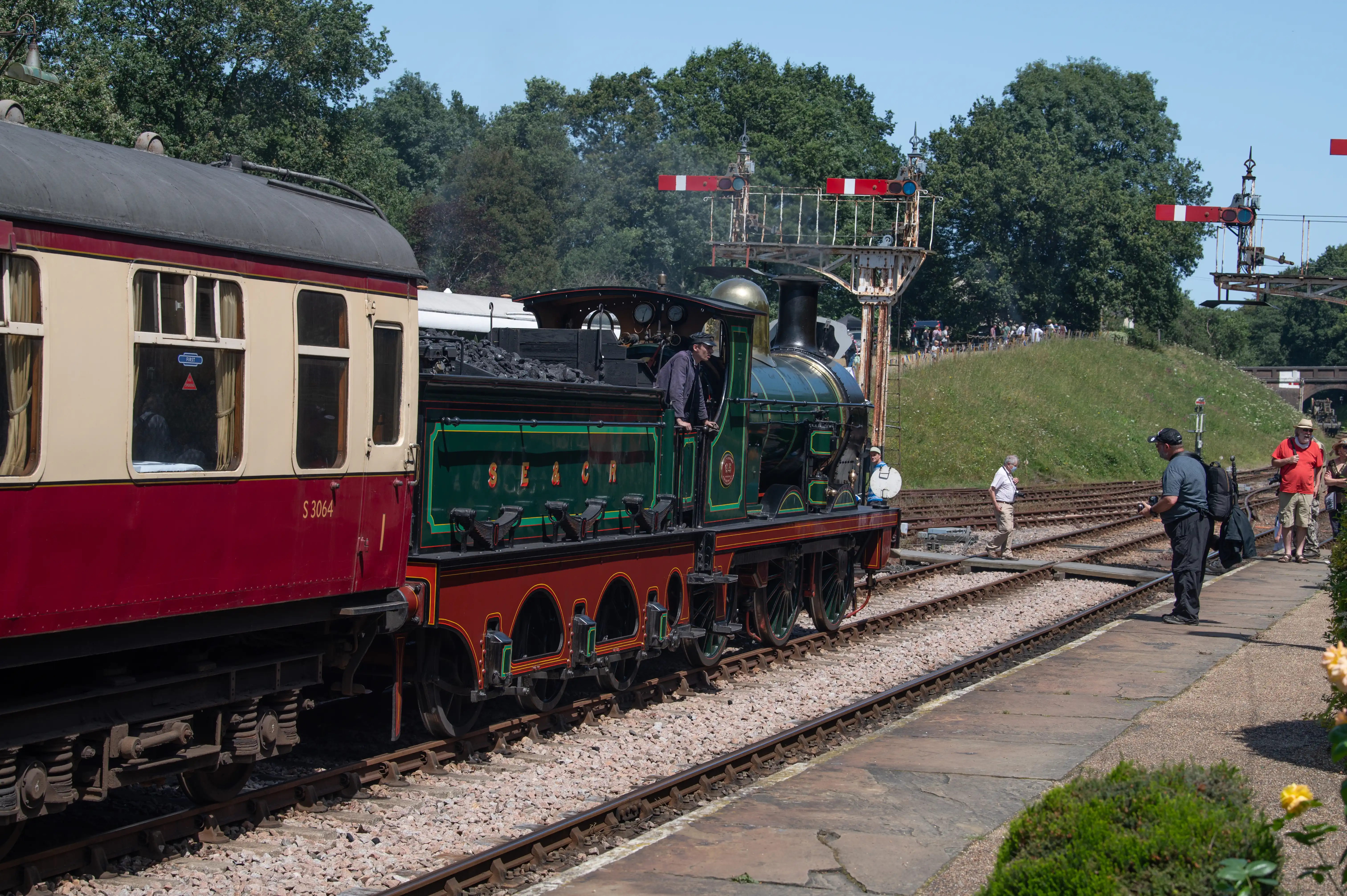 A side view of a vintage green and red steam train, numbered S 3064, alongside a maroon and cream carriage at a railway station. People are visible on the platform, with railway signals and trees in the background.