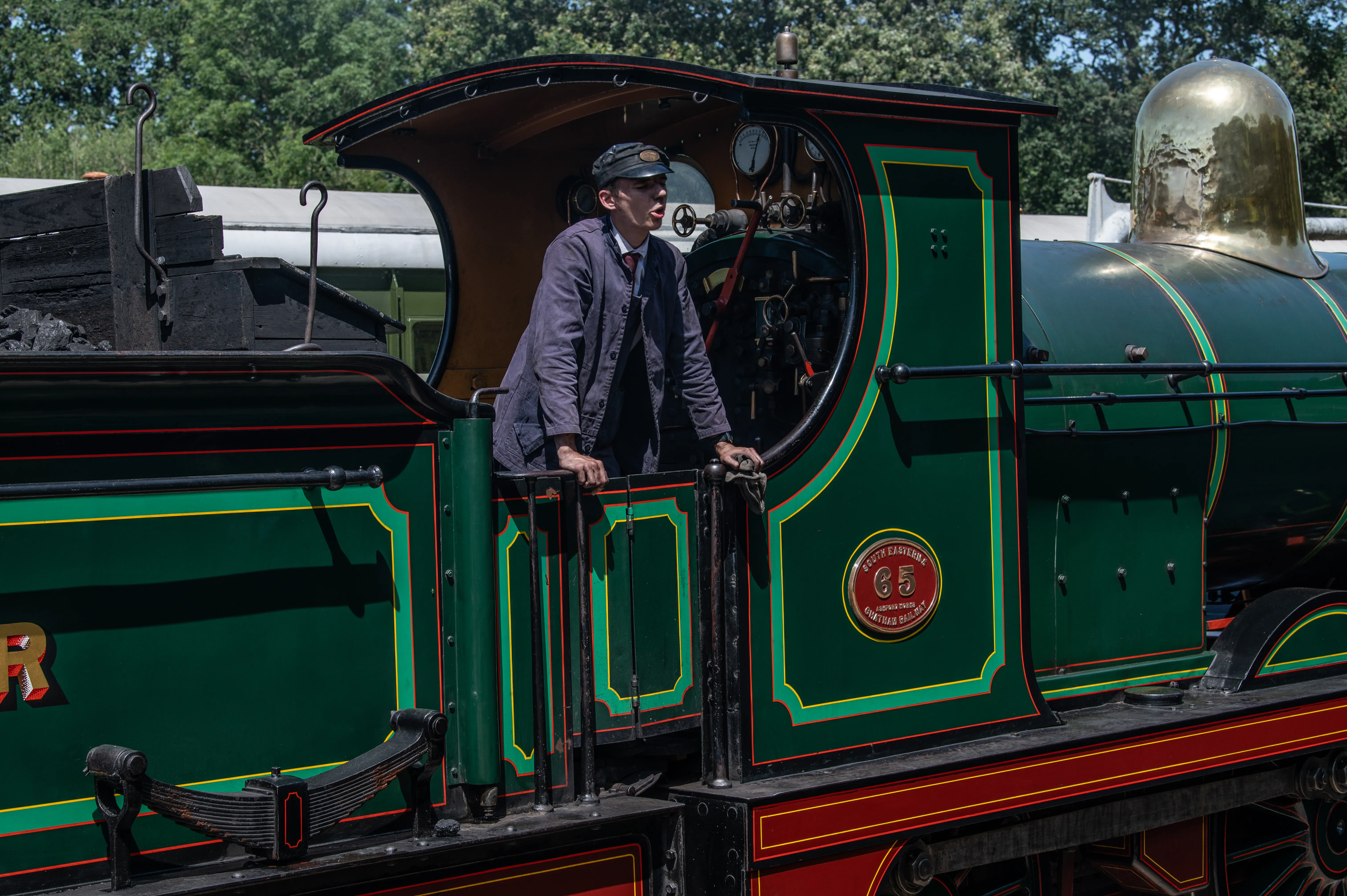 A man in a vintage train driver's uniform stands in the cab of a steam locomotive, with his hands on the railing. The locomotive is green with red and gold detailing and has a round emblem on its side.