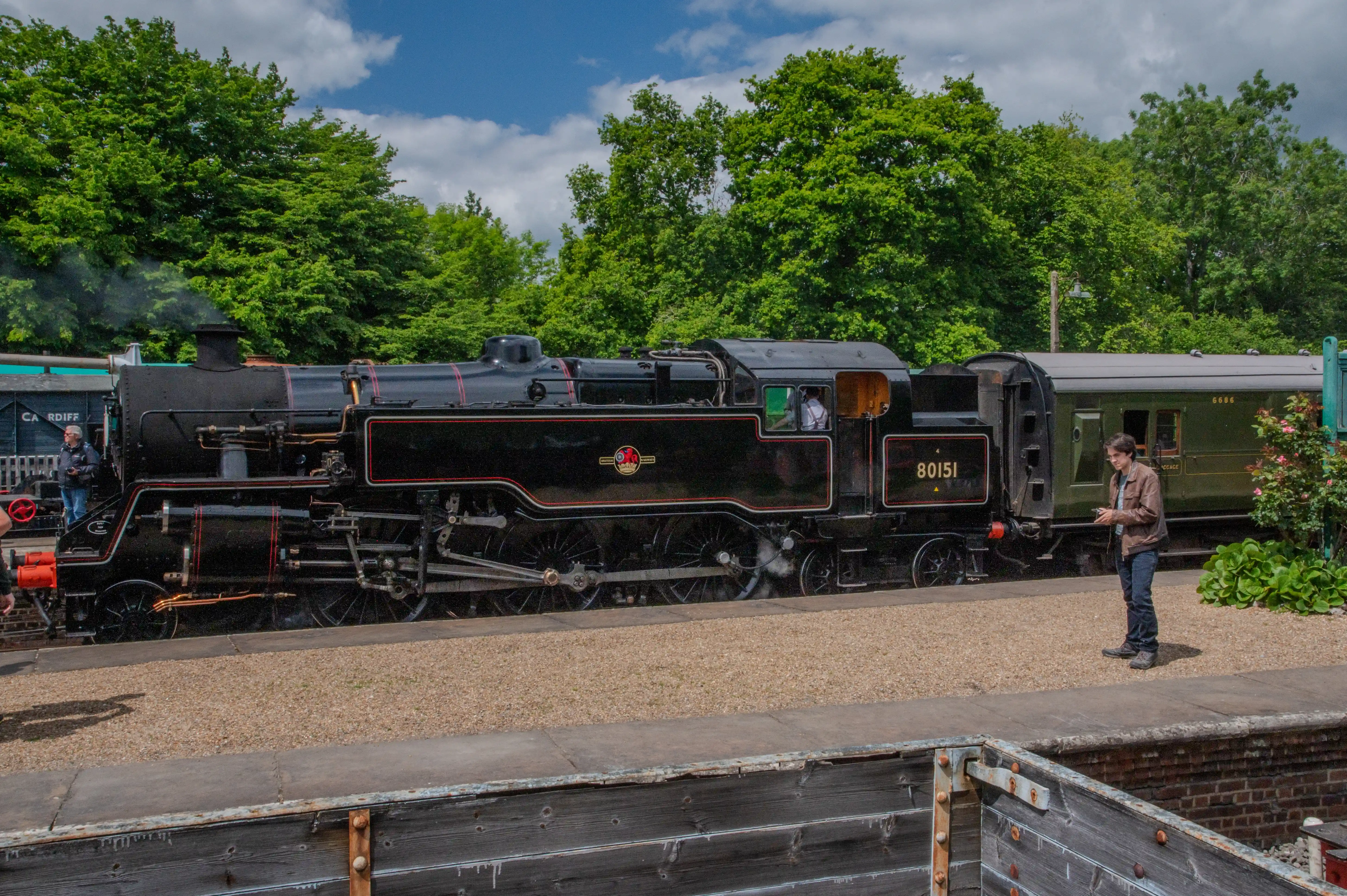 A black steam locomotive numbered 80151 is at a station platform. A young man in a brown jacket stands nearby, looking at his mobile phone. Trees and a blue sky with clouds are in the background.