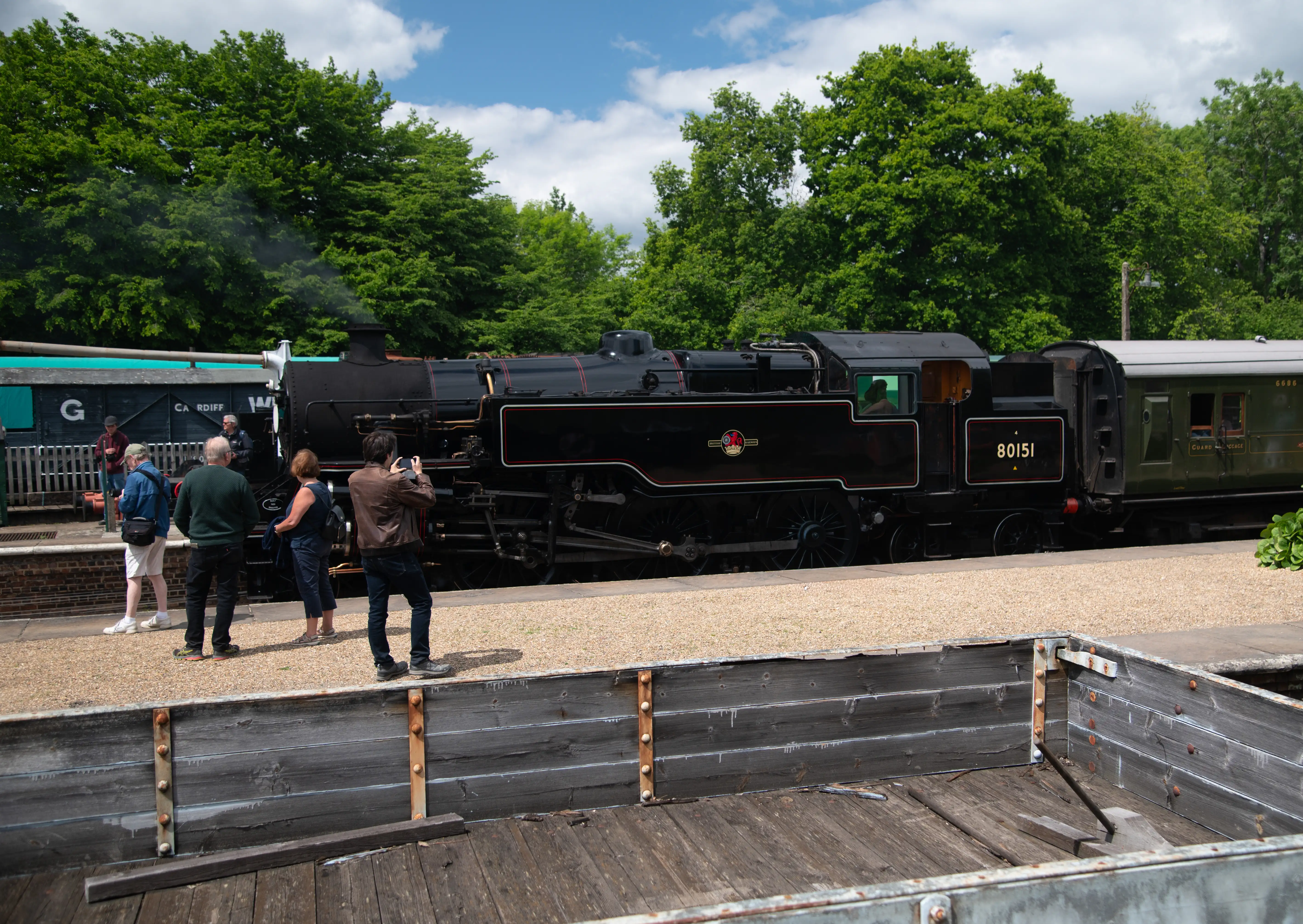 A black steam train with the number 80151 on its side is stationary at a station platform. Several people are standing on the platform, one of whom is taking a photograph of the train. Green trees line the background, and a blue sky with white clouds is overhead. A rustic wooden structure is in the foreground.