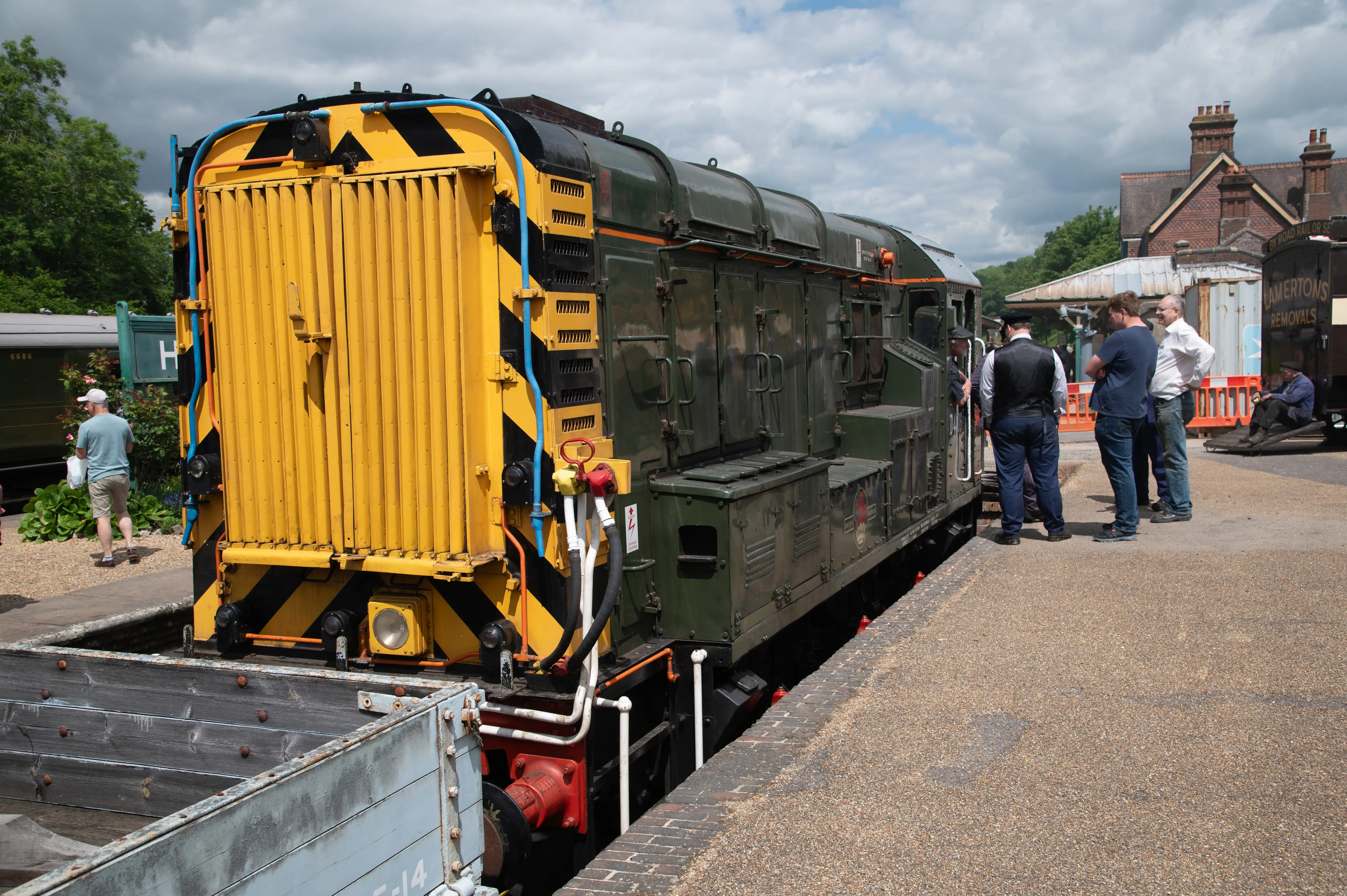 Dark-green diesel locomotive with yellow-and-black warning stripes at a heritage railway platform, with people standing beside it.