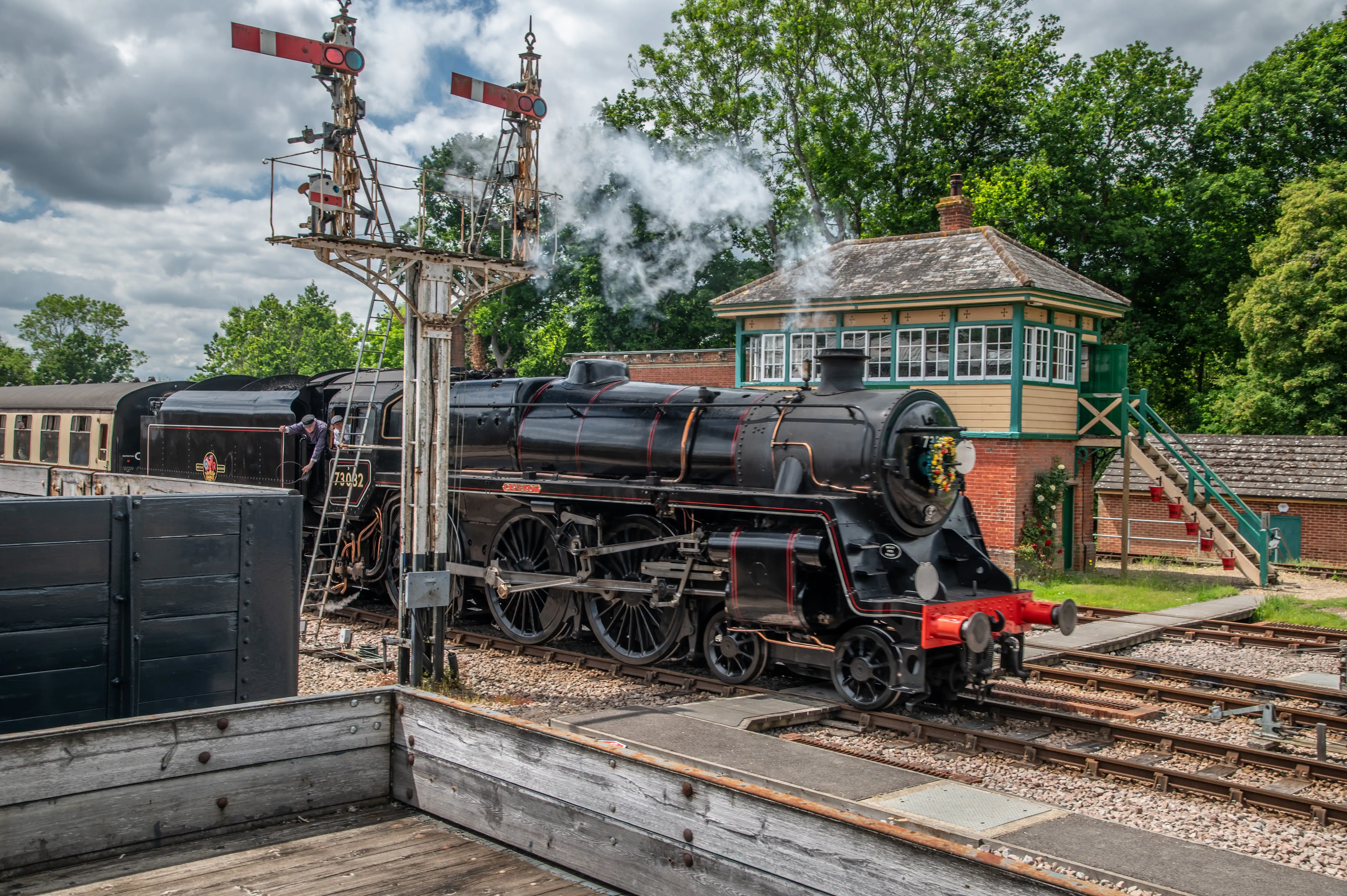 A black steam train with red trim, numbered 73032, emits steam next to a quaint green and yellow signal box. Railway signals are positioned above the train.