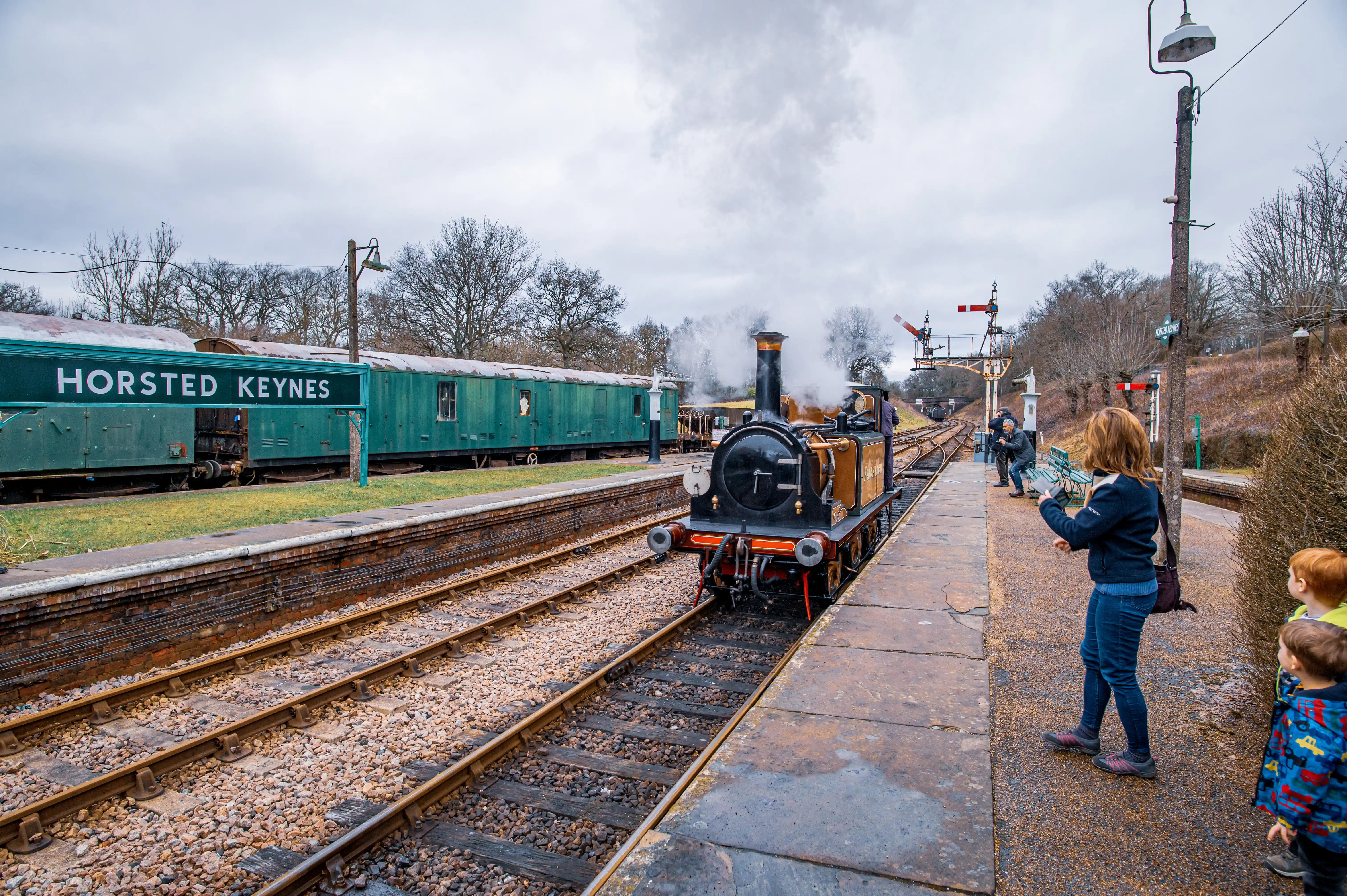 Steam locomotive arriving at Horsted Keynes station with passengers watching from the platform.