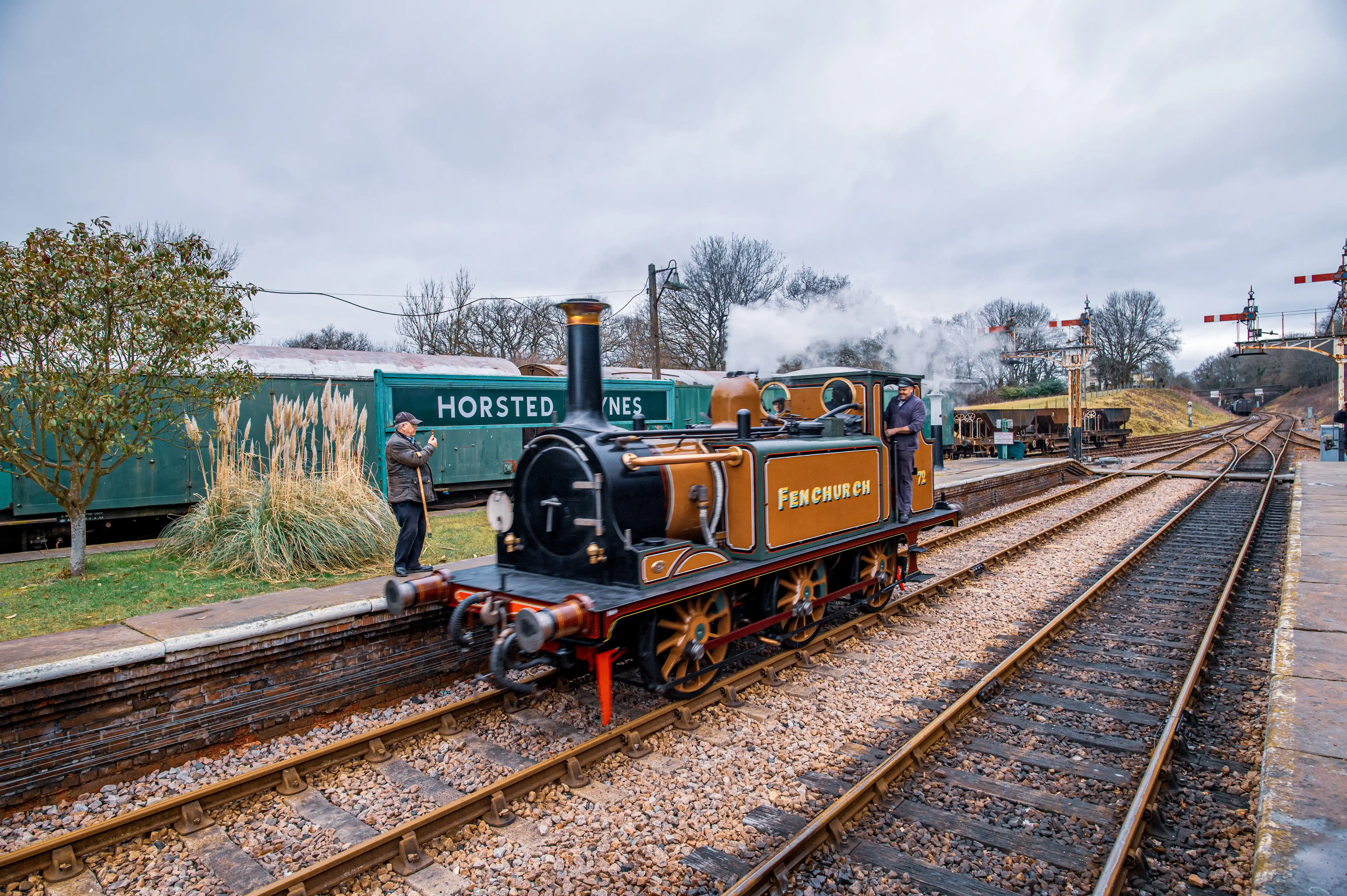A small, orange and black steam engine with Fenchurch written on its side is parked on railway tracks at Horsted Keynes station. Steam billows from its chimney. A man in a brown jacket and cap is standing on the platform to the left, holding a camera and a stick. Another man in a dark coat stands on the platform to the right. Green train carriages are behind the locomotive, with a sign reading HORSTED KEYNES. The sky is overcast.