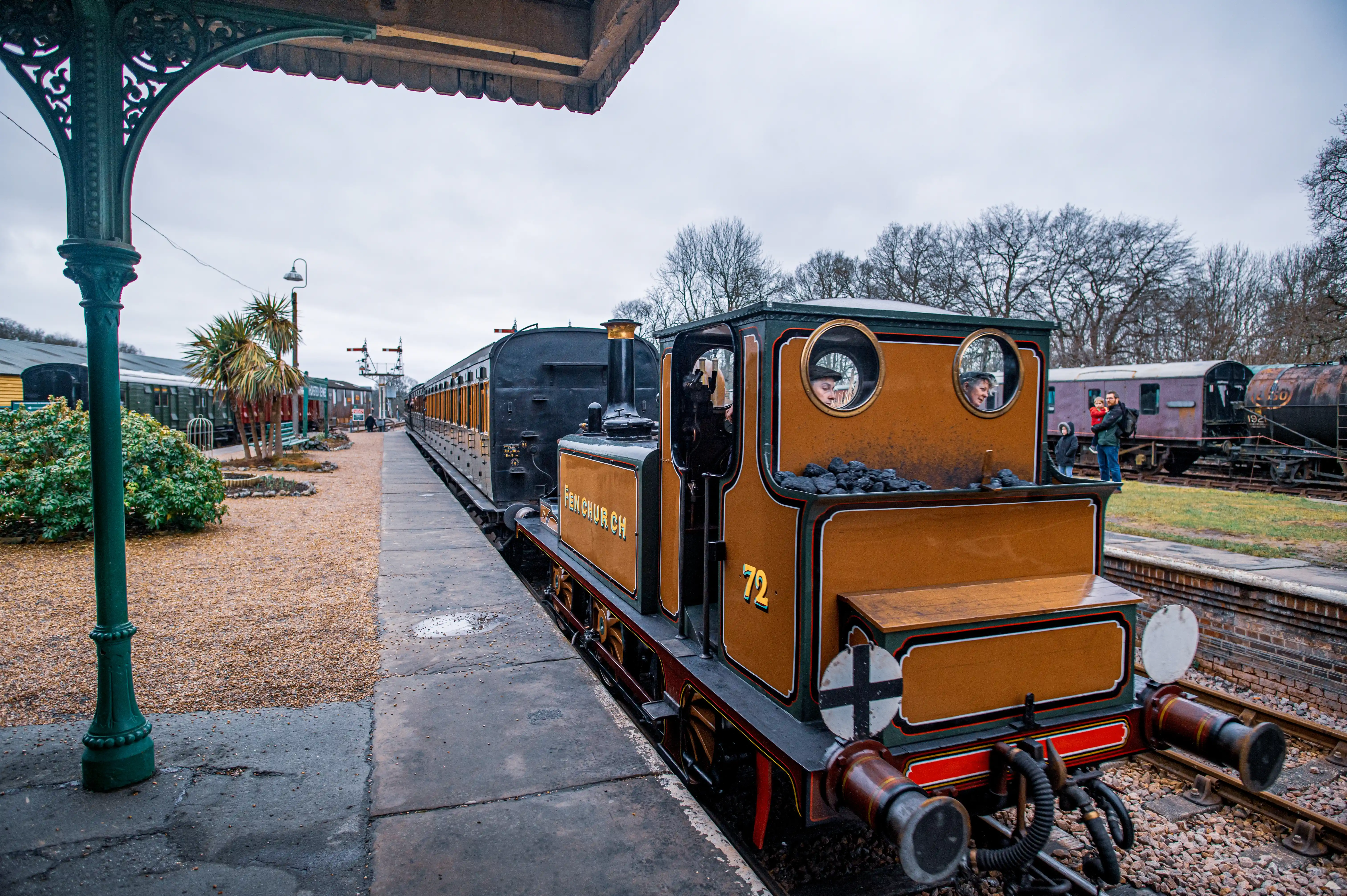 A close-up view of a miniature steam locomotive named Fenchurch, numbered 72, positioned on a railway platform. The train is predominantly orange with green trim. Several people, including a driver and passengers, are visible within and around the locomotive. In the background, other train carriages and people can be seen, along with trees and a cloudy sky.
