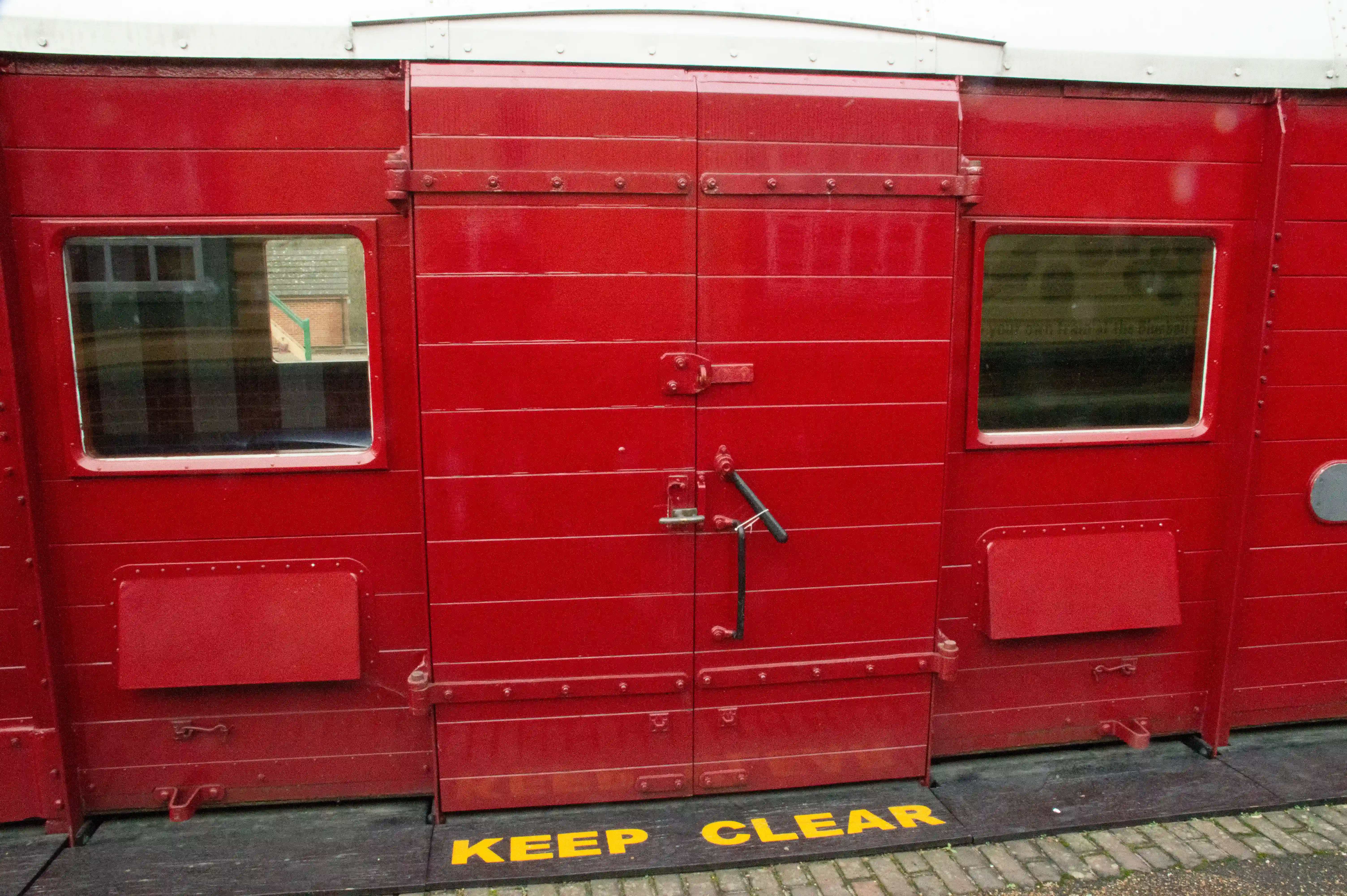Side view of a bright red train carriage, focusing on the double doors and two windows. A yellow KEEP CLEAR warning is visible on the ground in the foreground.