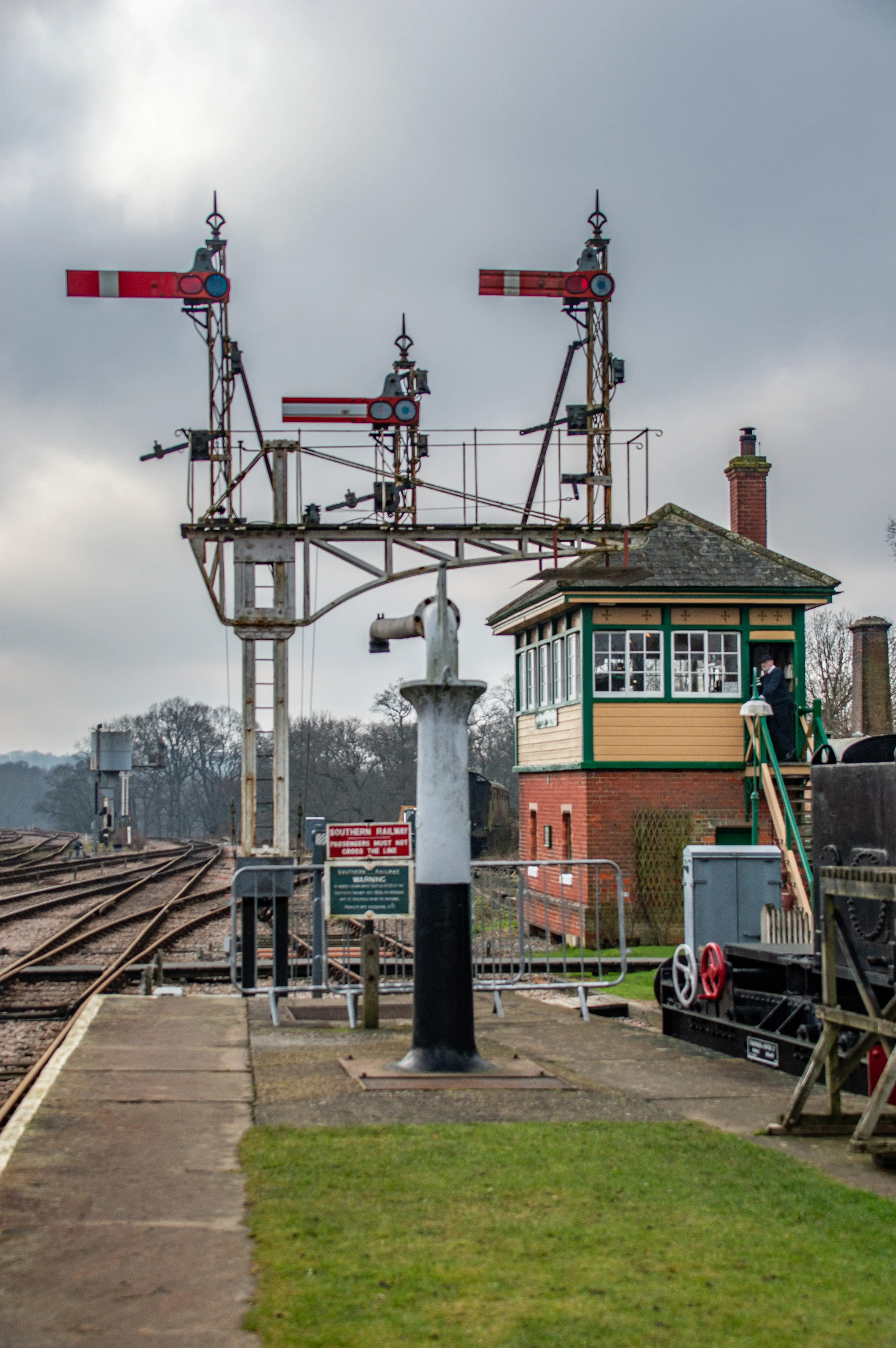 Two classic railway semaphore signals are positioned above a brick and timber signal box, a man in vintage attire stands on the signal box steps. Railway tracks spread out in the foreground under a cloudy sky.