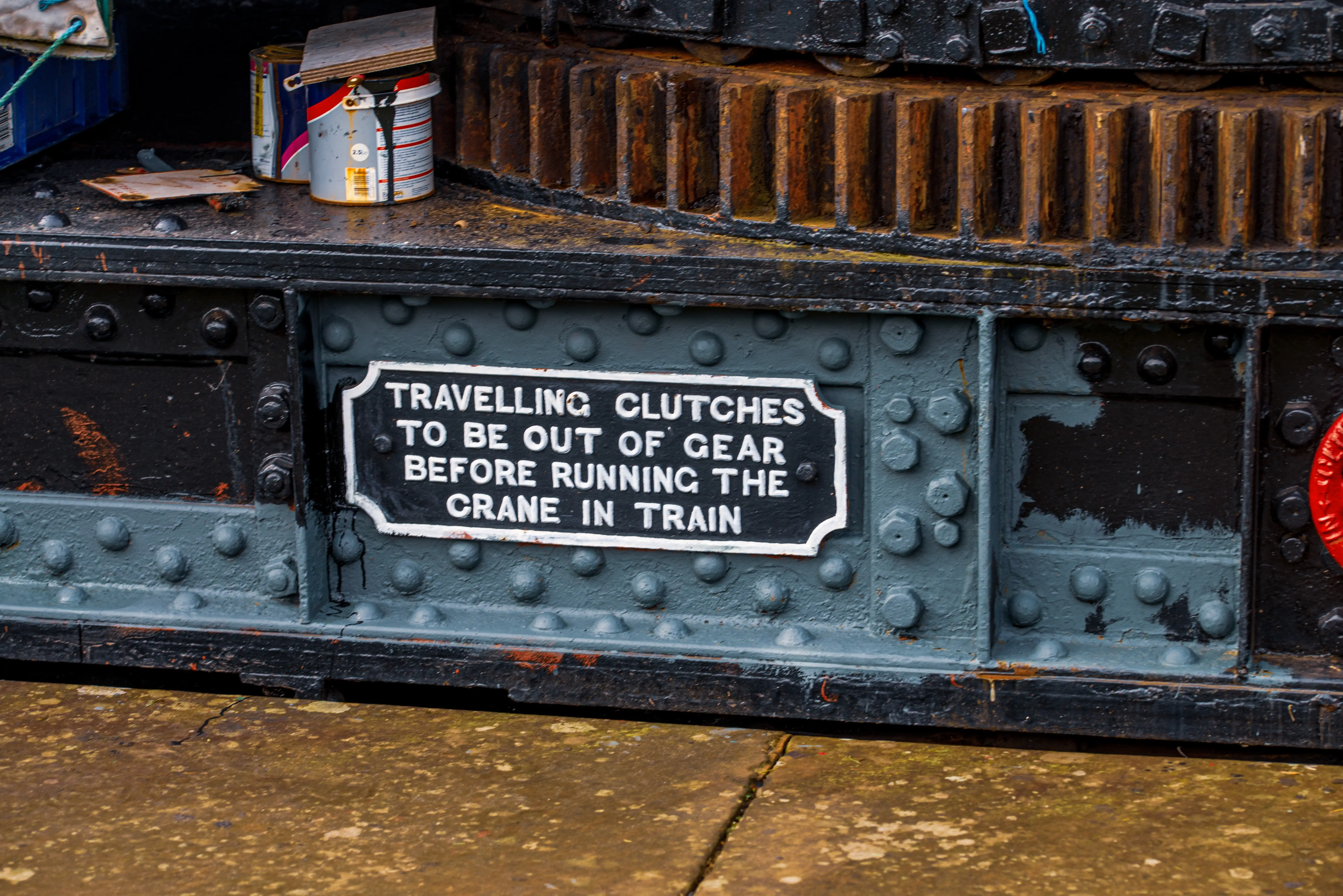 A black and white warning sign with the text TRAVELLING CLUTCHES TO BE OUT OF GEAR BEFORE RUNNING THE CRANE IN TRAIN
