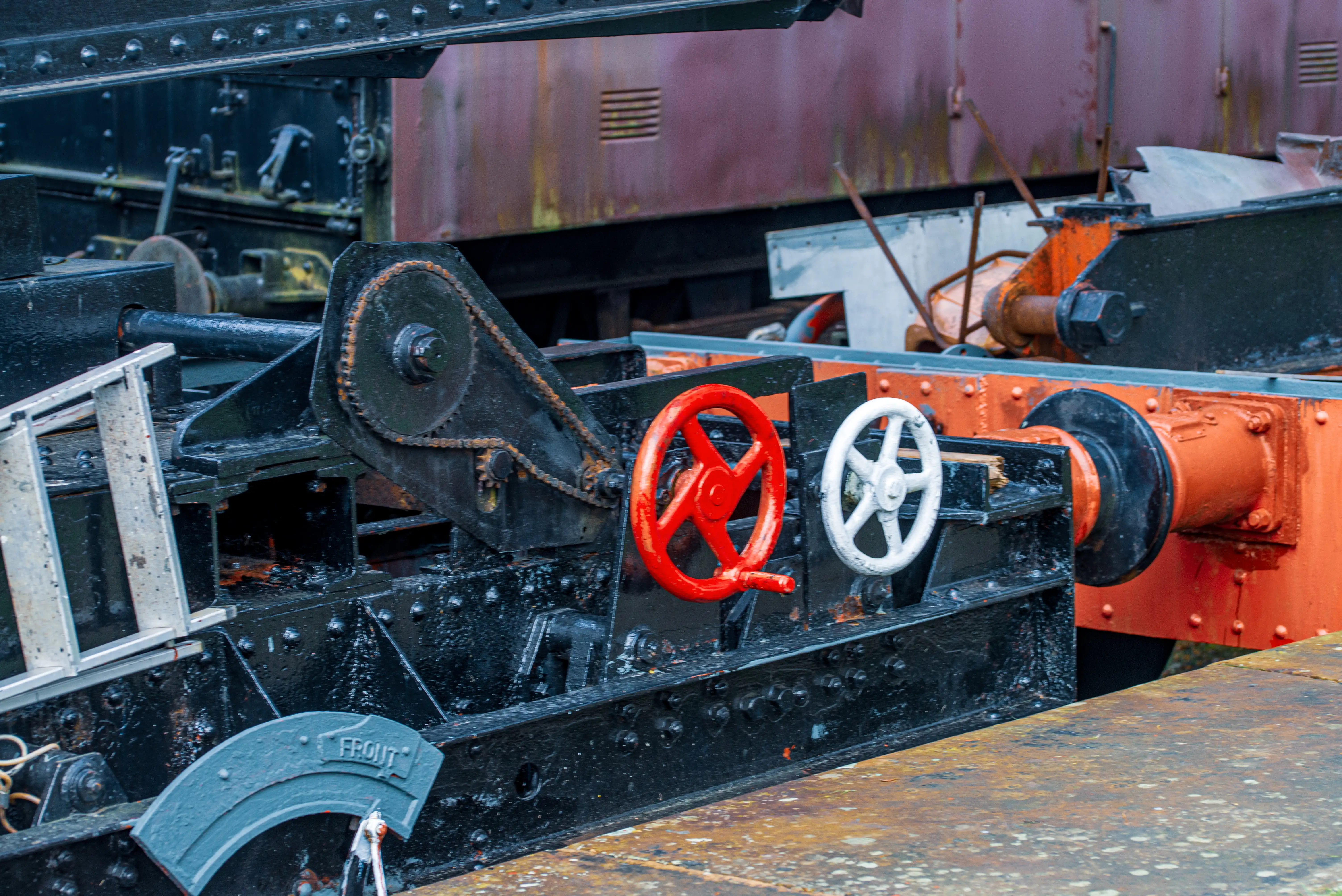 Close-up of a mechanical assembly on a vintage train, featuring a chain drive, a red handwheel, and a white handwheel, against a backdrop of weathered metal and orange painted components.