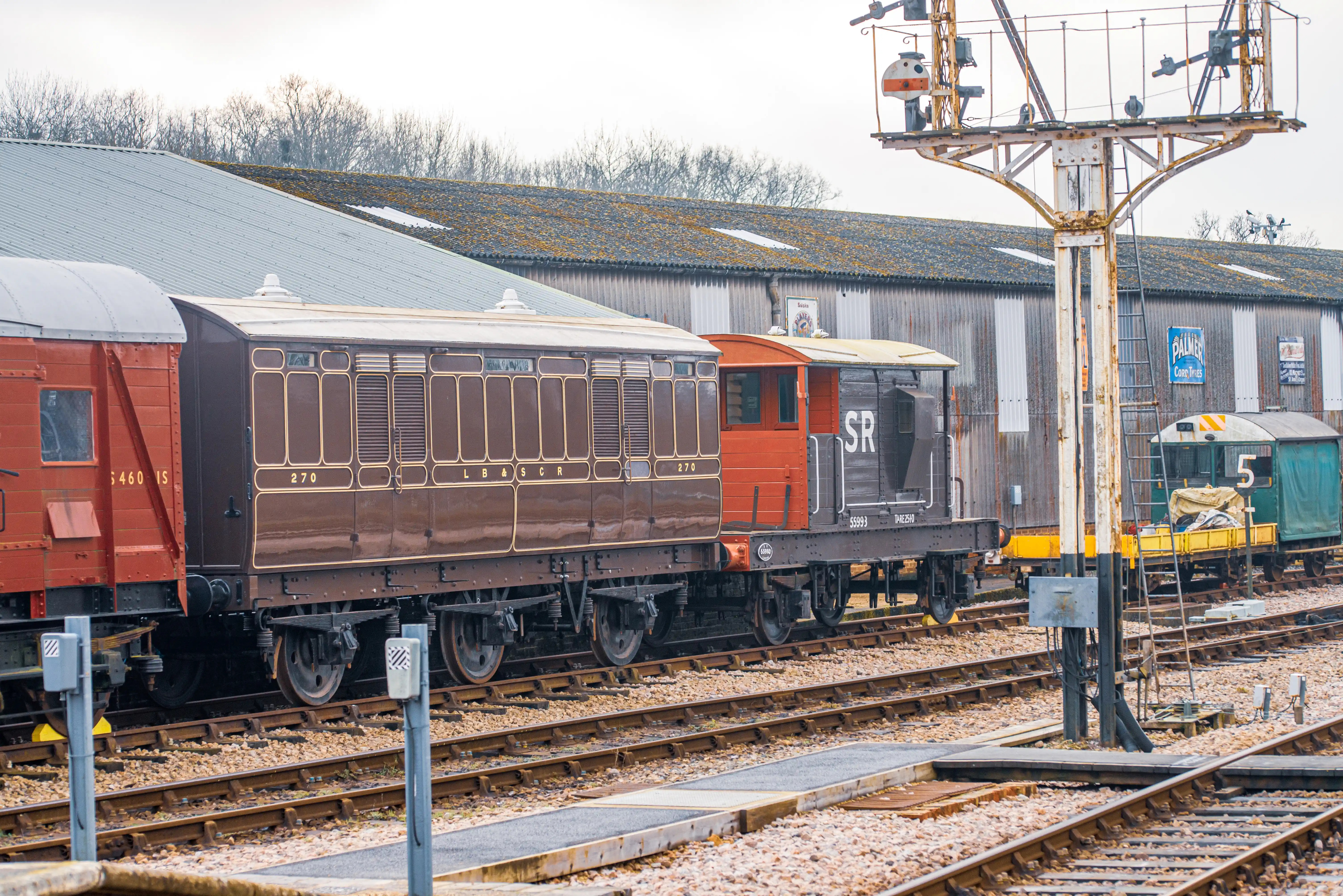 Multiple historic train carriages, one brown with detailed windows and the number 270, and a red goods wagon, are stationary on railway tracks. Beside them is a dark locomotive with 