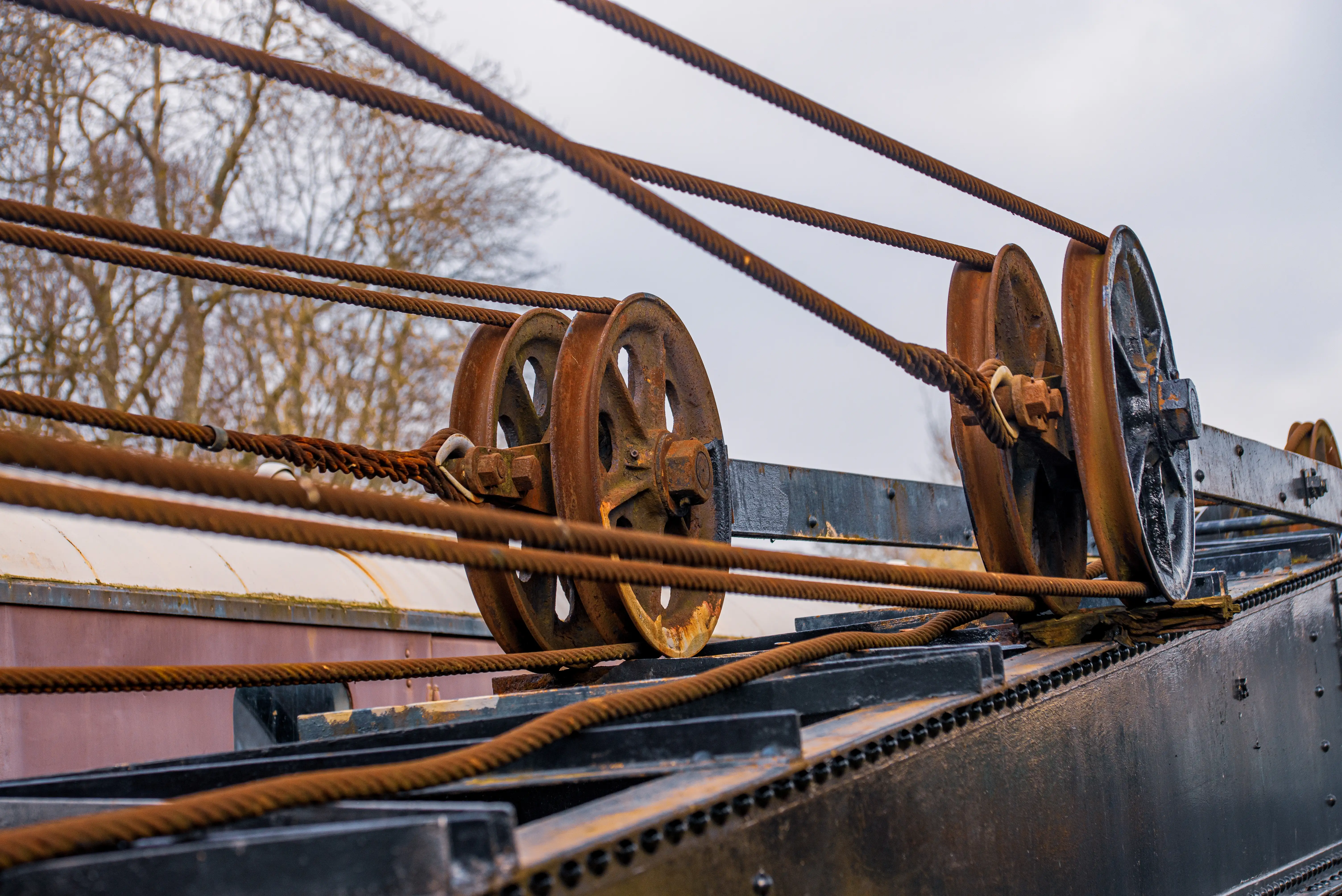 Rusted metal pulleys with steel cables mounted on an industrial frame outdoors with winter trees behind.