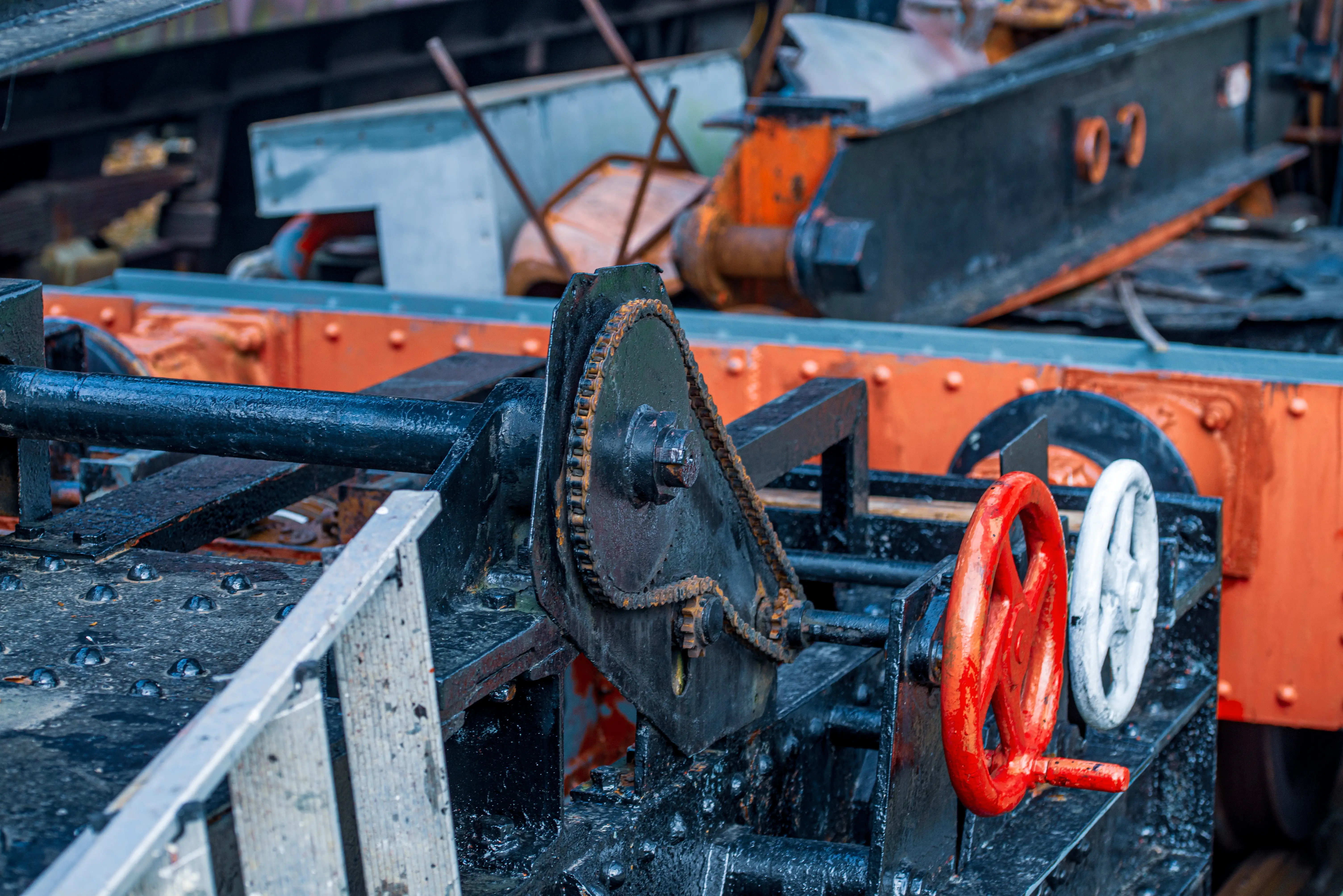 Close-up of the rusted chain drive and red and white hand wheels on the side of a vintage train carriage, with bolts and a metal ladder visible.