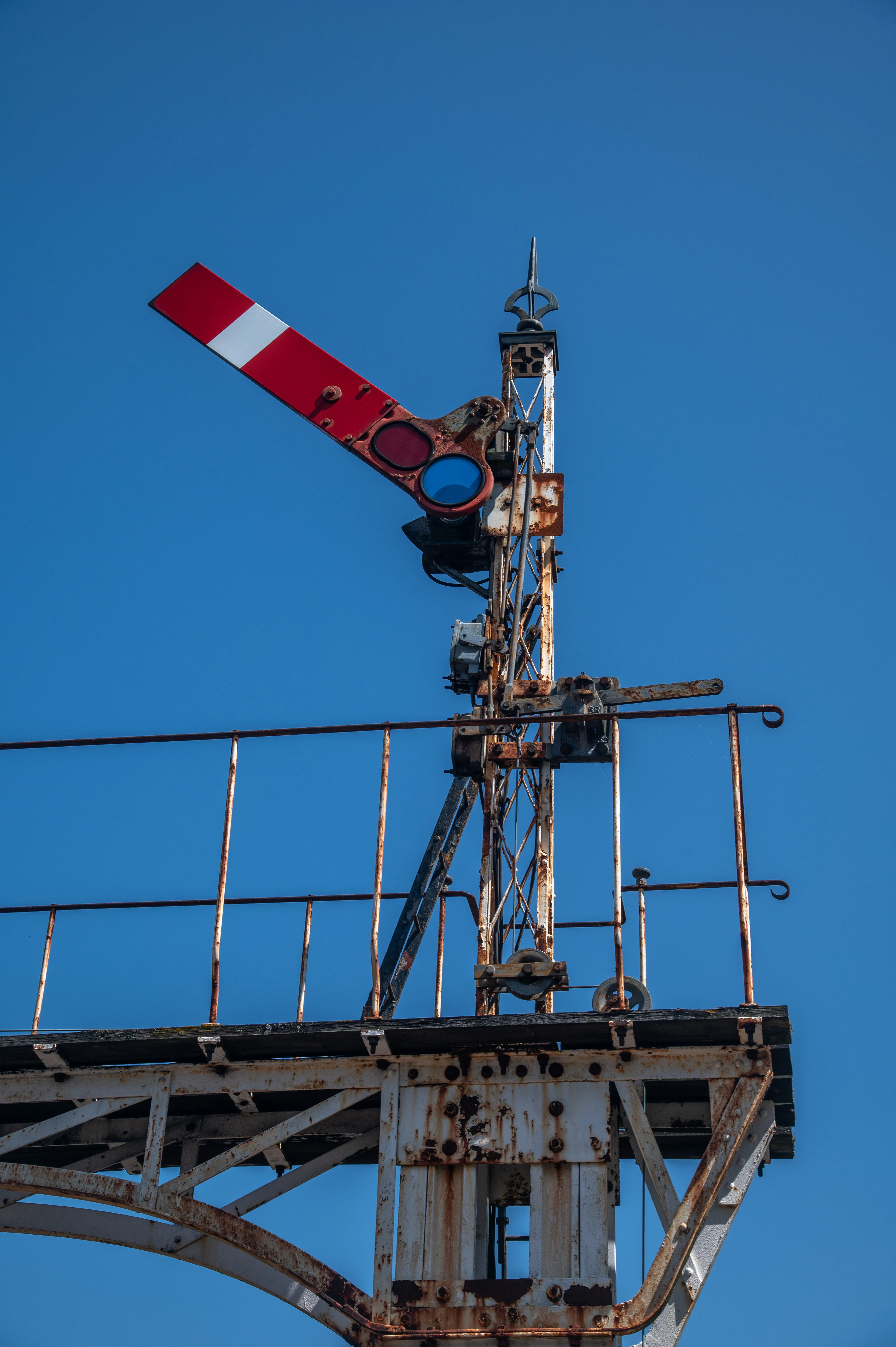 A close-up of a rusty, vintage railway signal with a red and white arm extended against a bright blue sky.