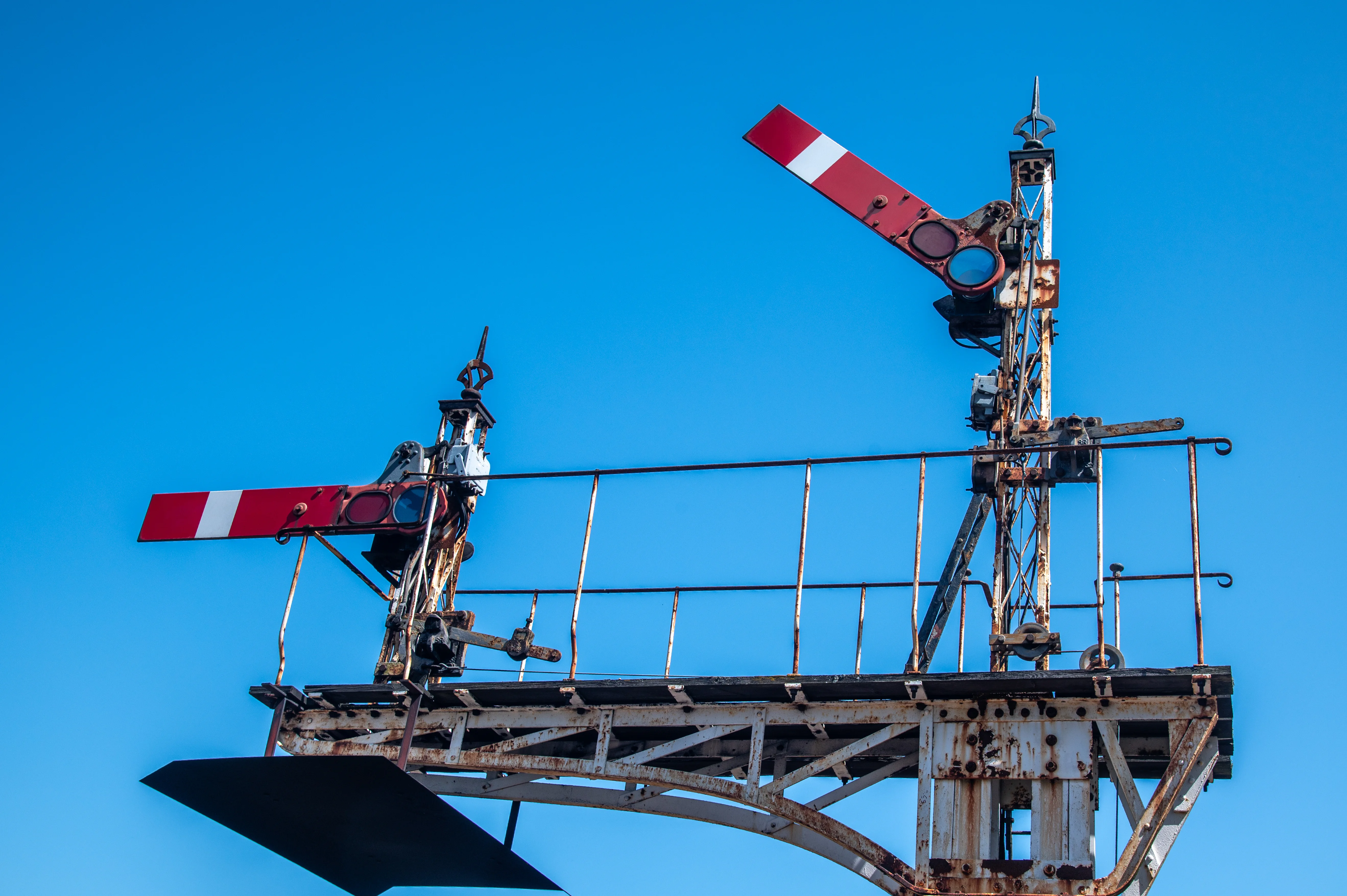 An image showing a pair of old, rusty railway semaphore signals, set against a bright blue sky. One signal arm points upwards and to the right, while the other points horizontally to the left. Both arms are painted with red and white stripes. The supporting structure is made of weathered metal.