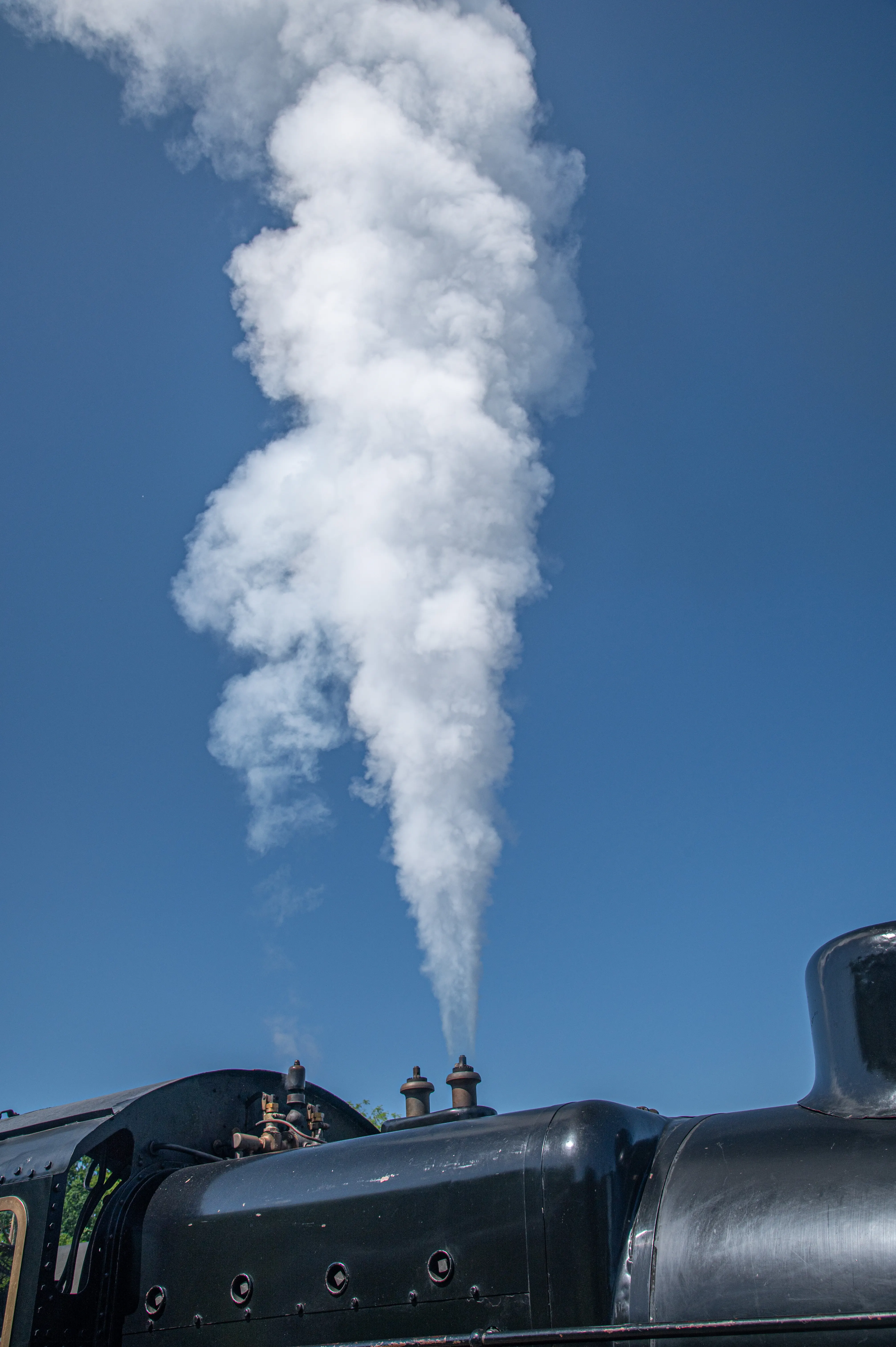 A close-up view of the top of a black steam locomotive with a large plume of white steam rising dramatically into a bright blue sky.