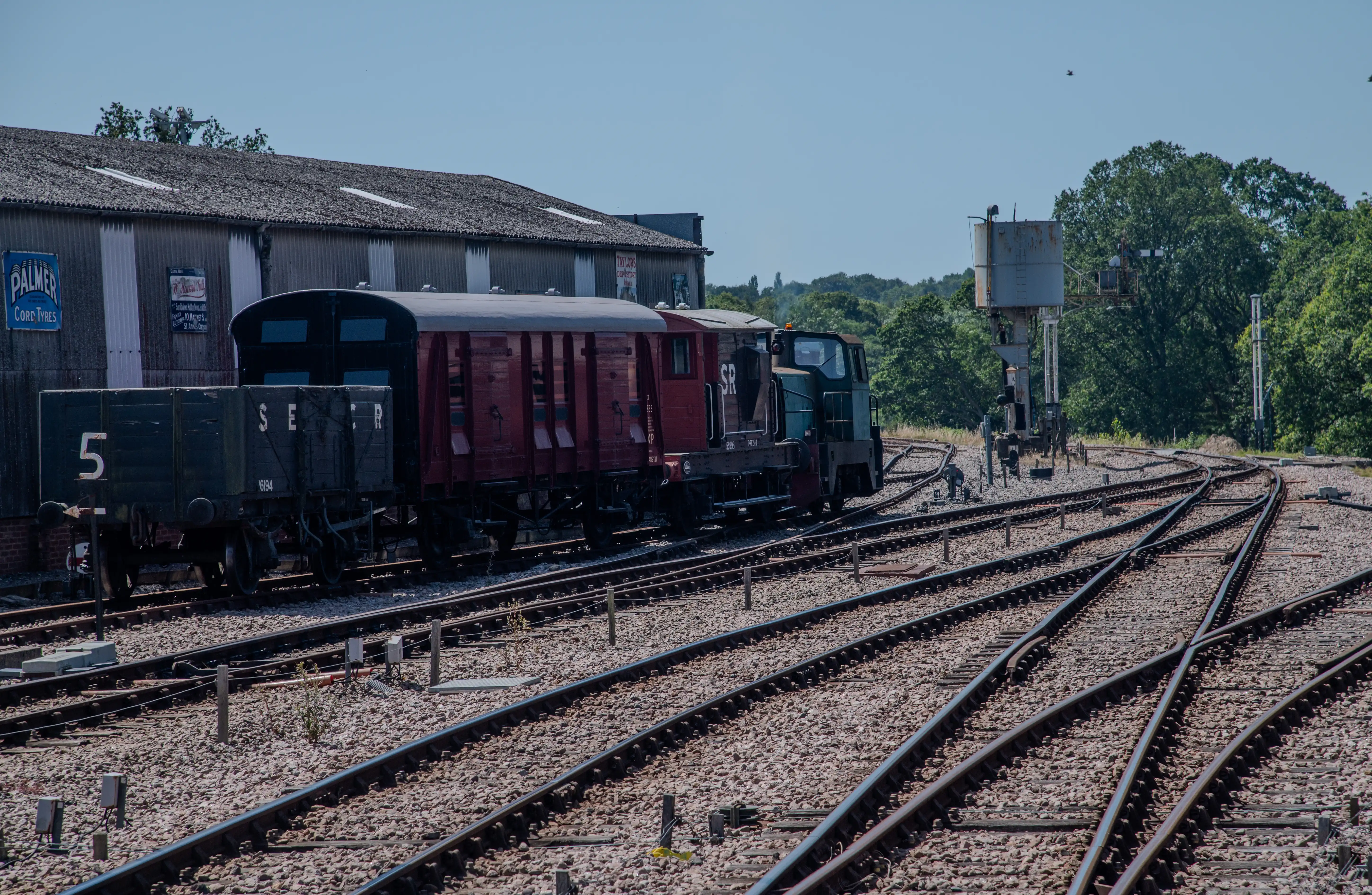 A line of vintage railway carriages and a locomotive are positioned on a set of curving railway tracks, with a corrugated iron building and greenery in the background.