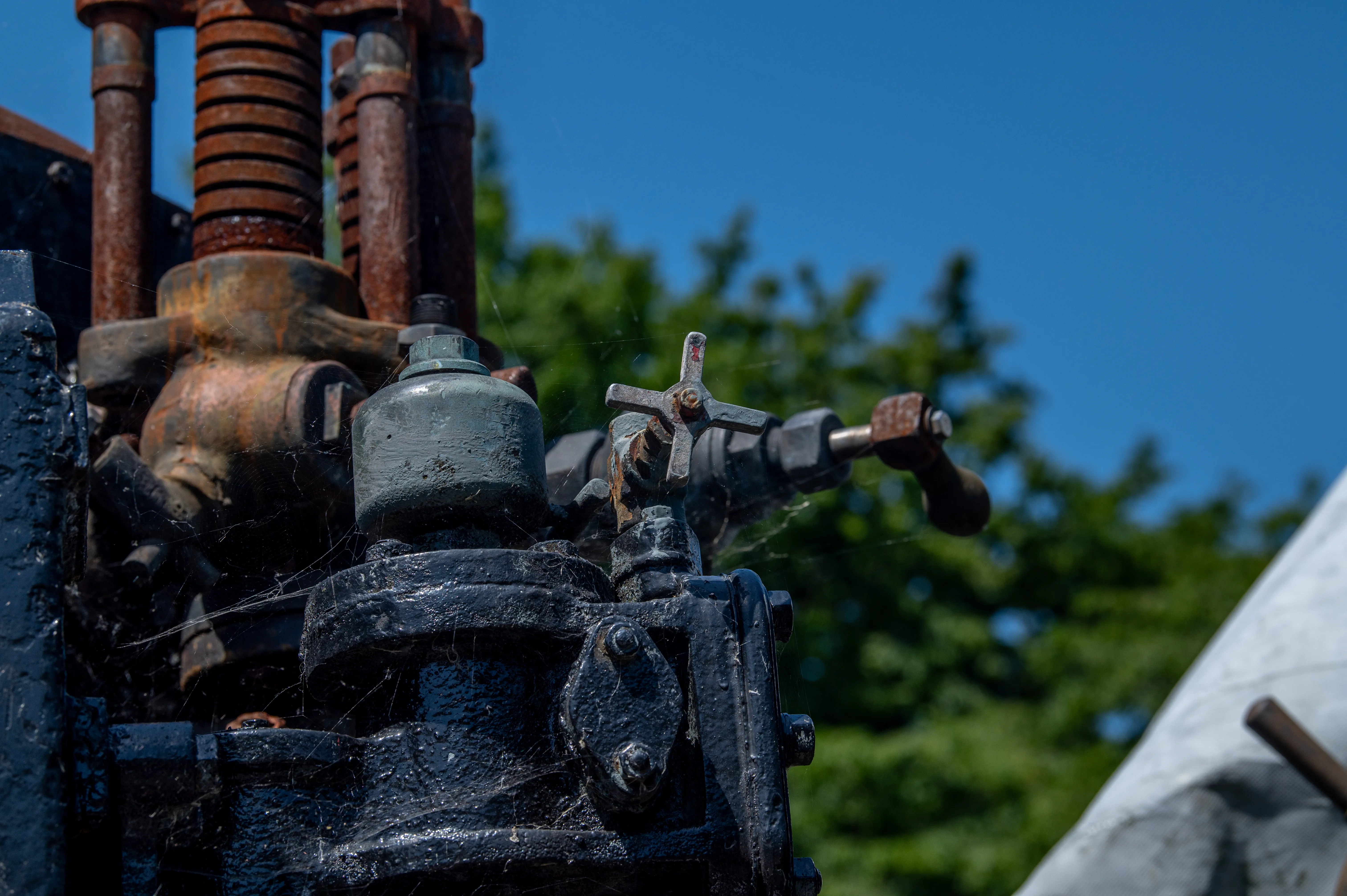 A close-up view of the intricate mechanical components of a vintage steam locomotive, including rusty metal pipes, a green-painted valve, a handwheel, and a cross-shaped lever. The background features a blurred blue sky and green foliage.