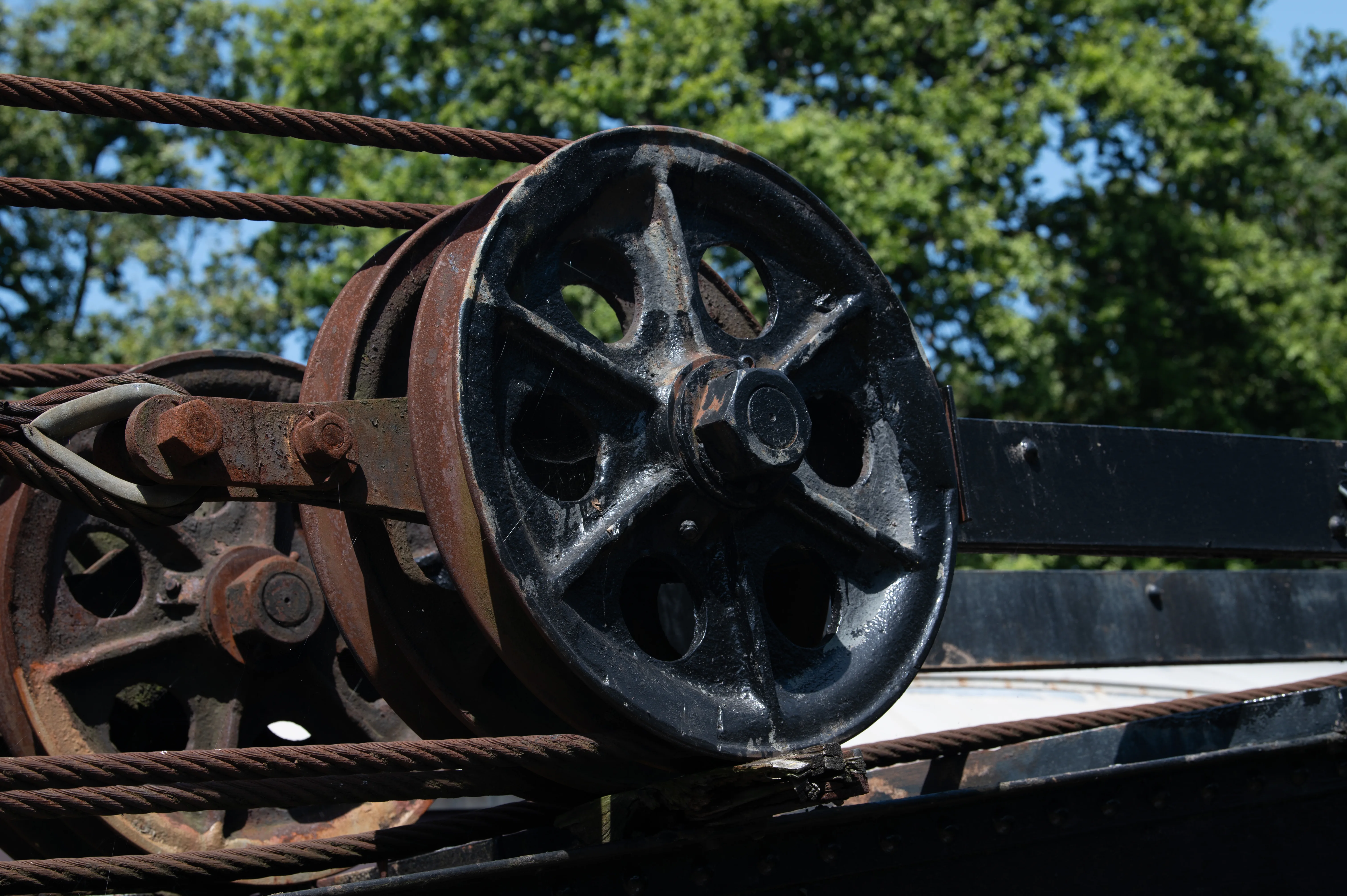 Close-up shot of an old, rusty pulley system with thick, weathered steel cables. One pulley is black and prominently featured, while another is partially visible and heavily rusted. Green foliage and a blue sky are out of focus in the background.
