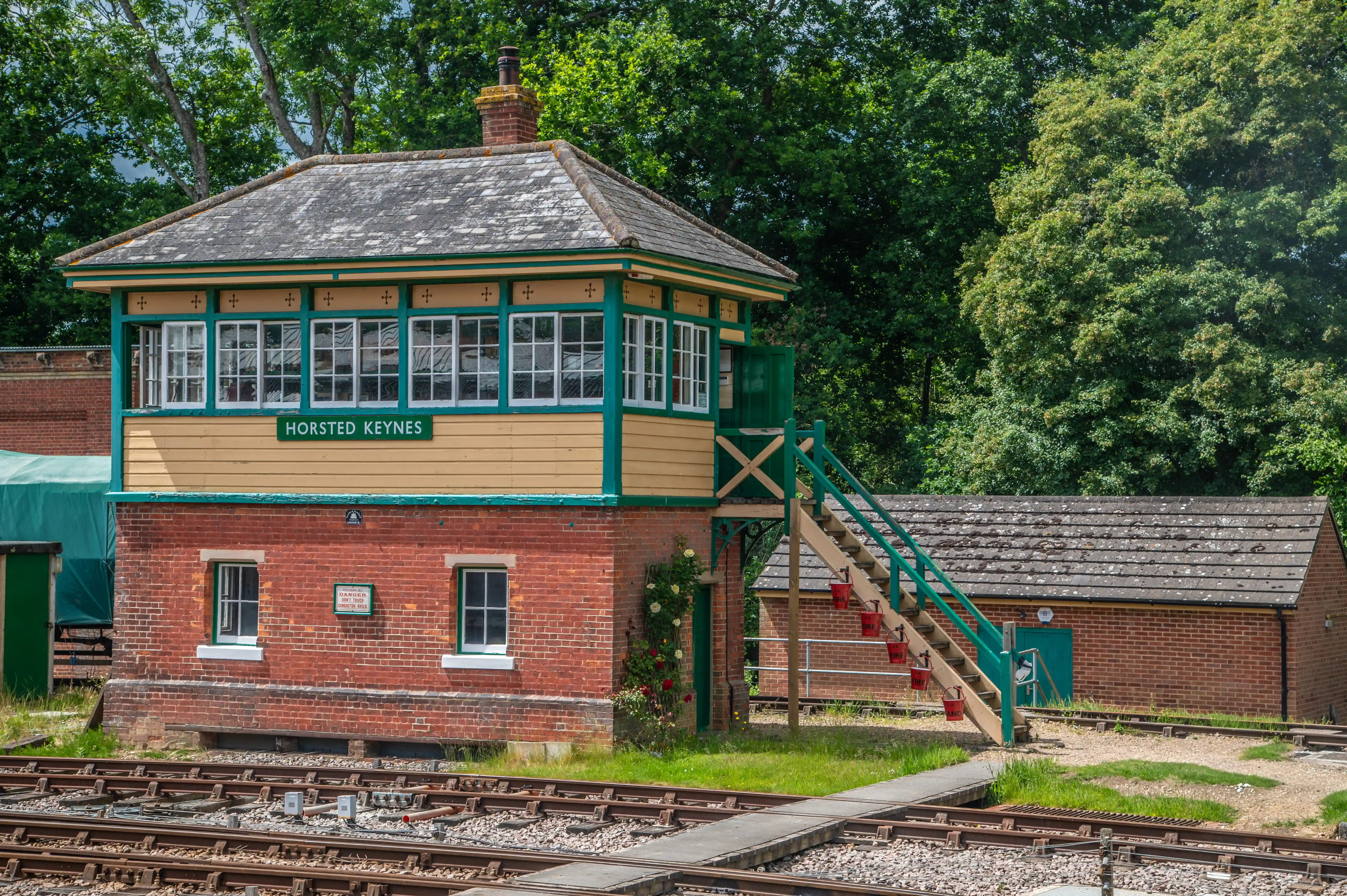A red brick signal box with a yellow and green upper section sits next to railway tracks, with dense green trees in the background.