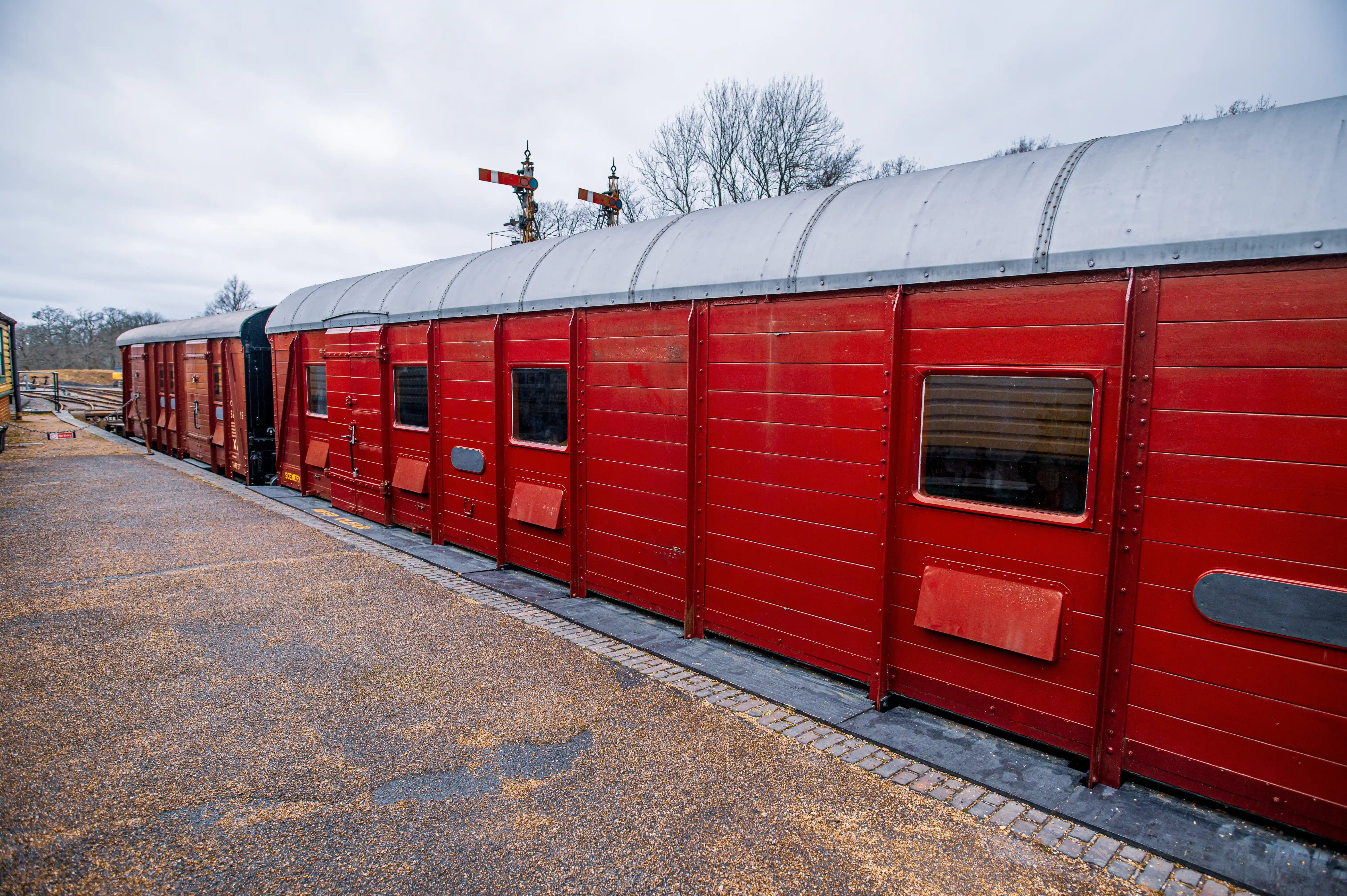 A row of classic red train carriages with silver curved roofs parked alongside a gravel and paved platform. Traditional railway signals stand tall in the background against a muted sky and bare trees.
