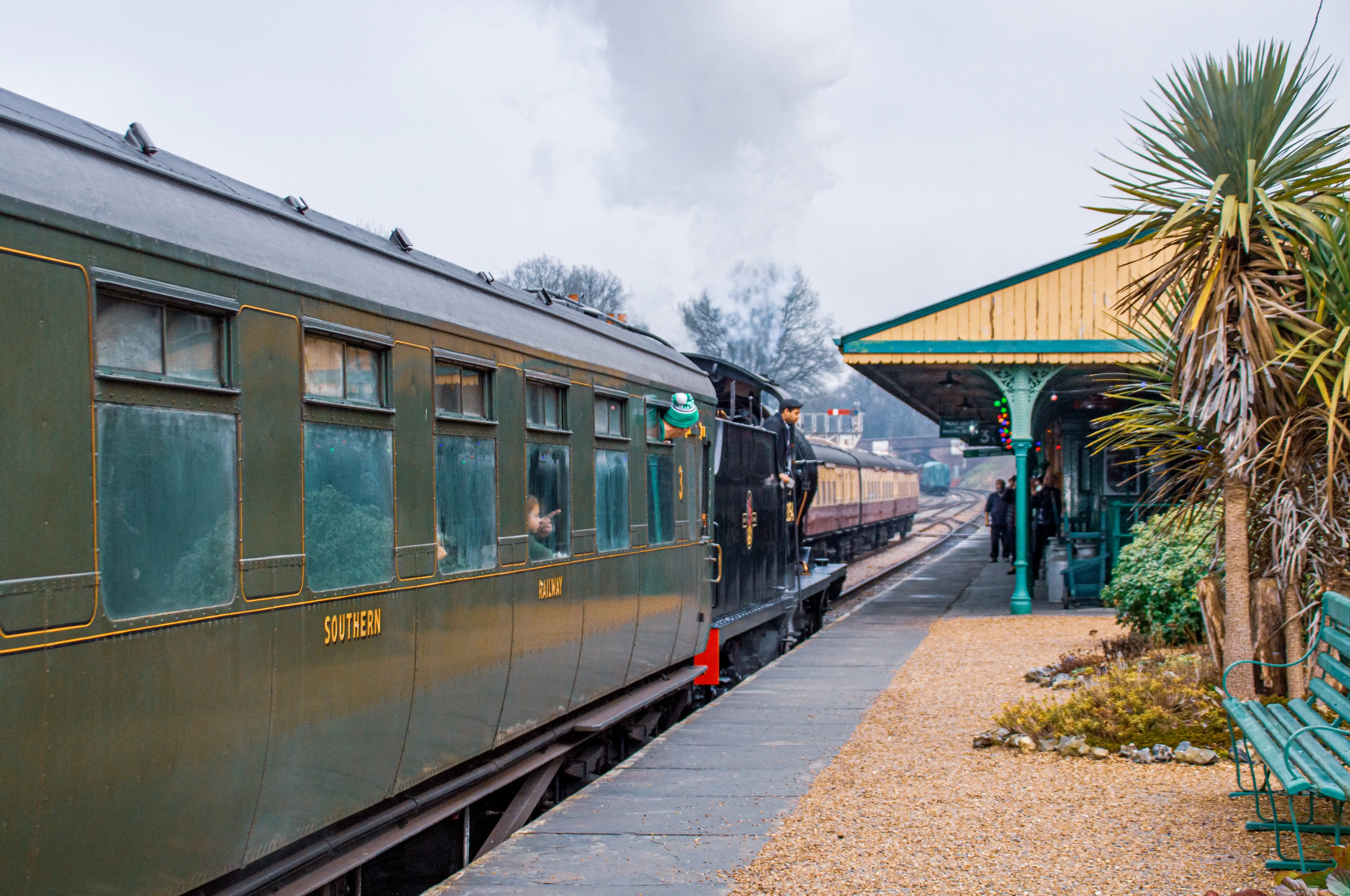 A dark green vintage train carriage with SOUTHERN written on the side, alongside a railway platform. In the background, a train locomotive and a station shelter are visible. A palm tree stands to the right.