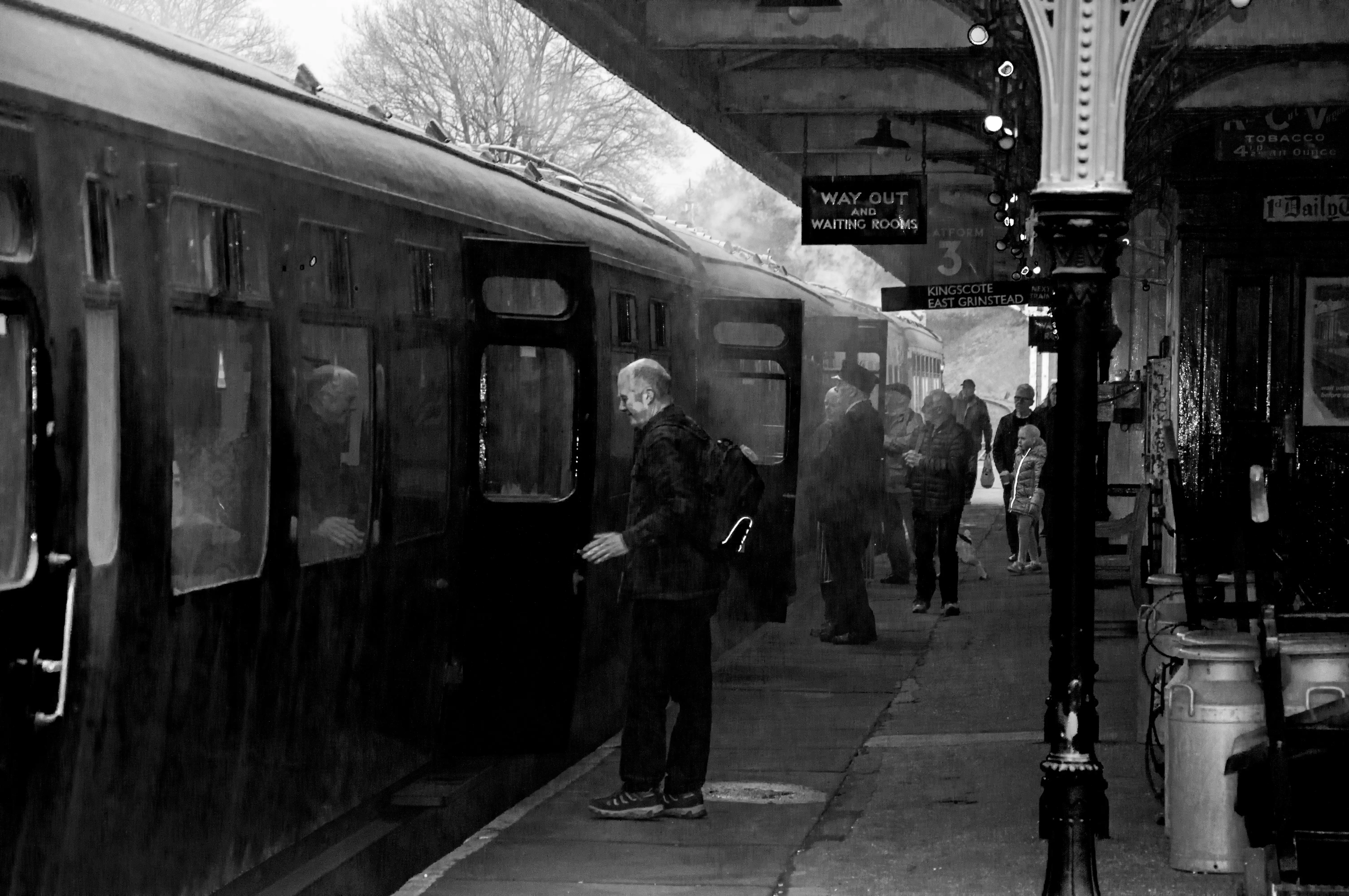 A black and white photograph of a vintage steam train at a platform. A man in a patterned jacket and dark trousers is stepping into the open door of a carriage. Several other people, some in hats and coats, are waiting on the platform. Signage indicates WAY OUT AND WAITING ROOMS and PLATFORM 3. Steam or smoke emanates from the train.