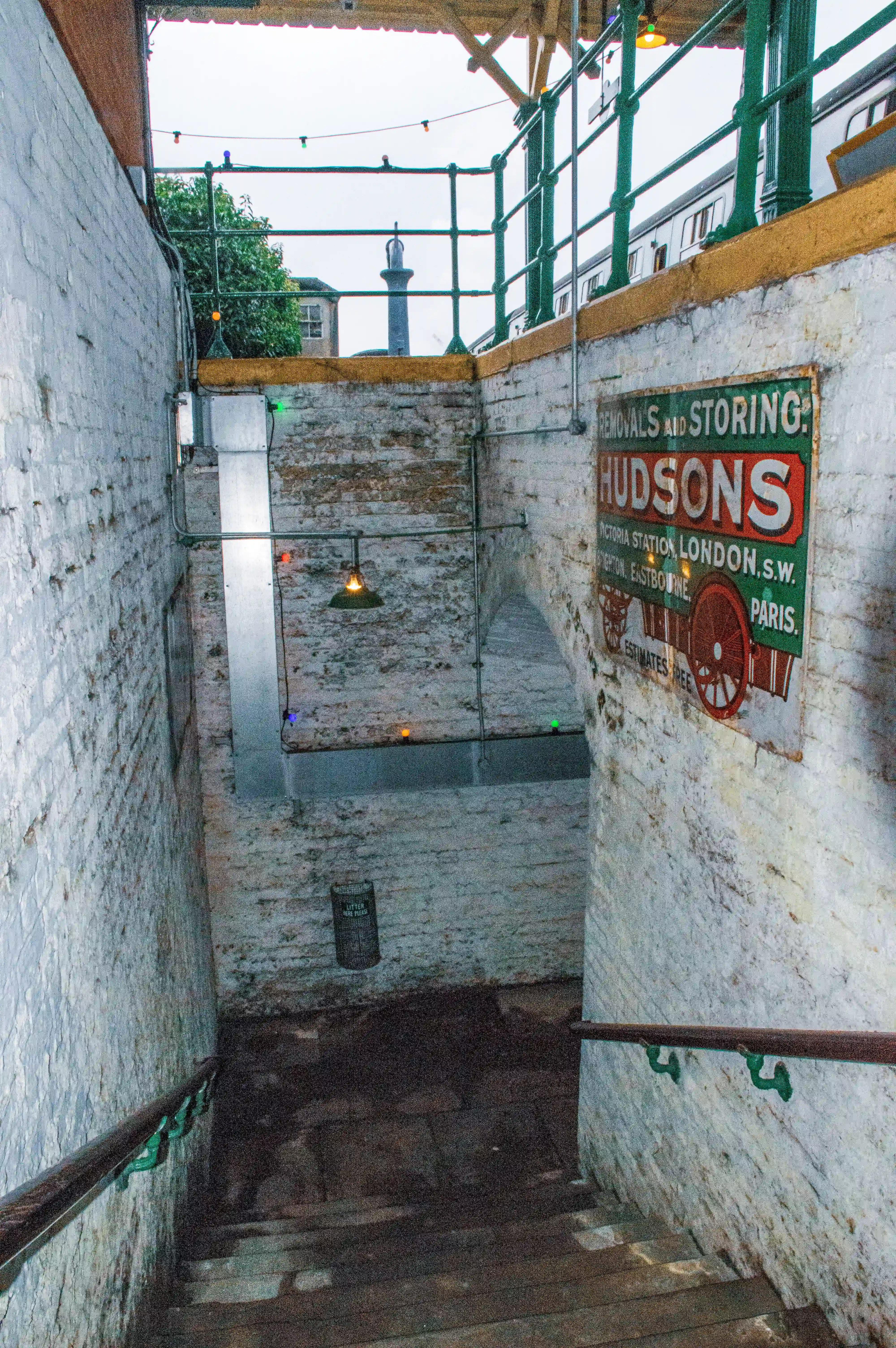 A view down stone steps at a railway station, with a vintage advert for Hudsons removals and storing on the right wall.