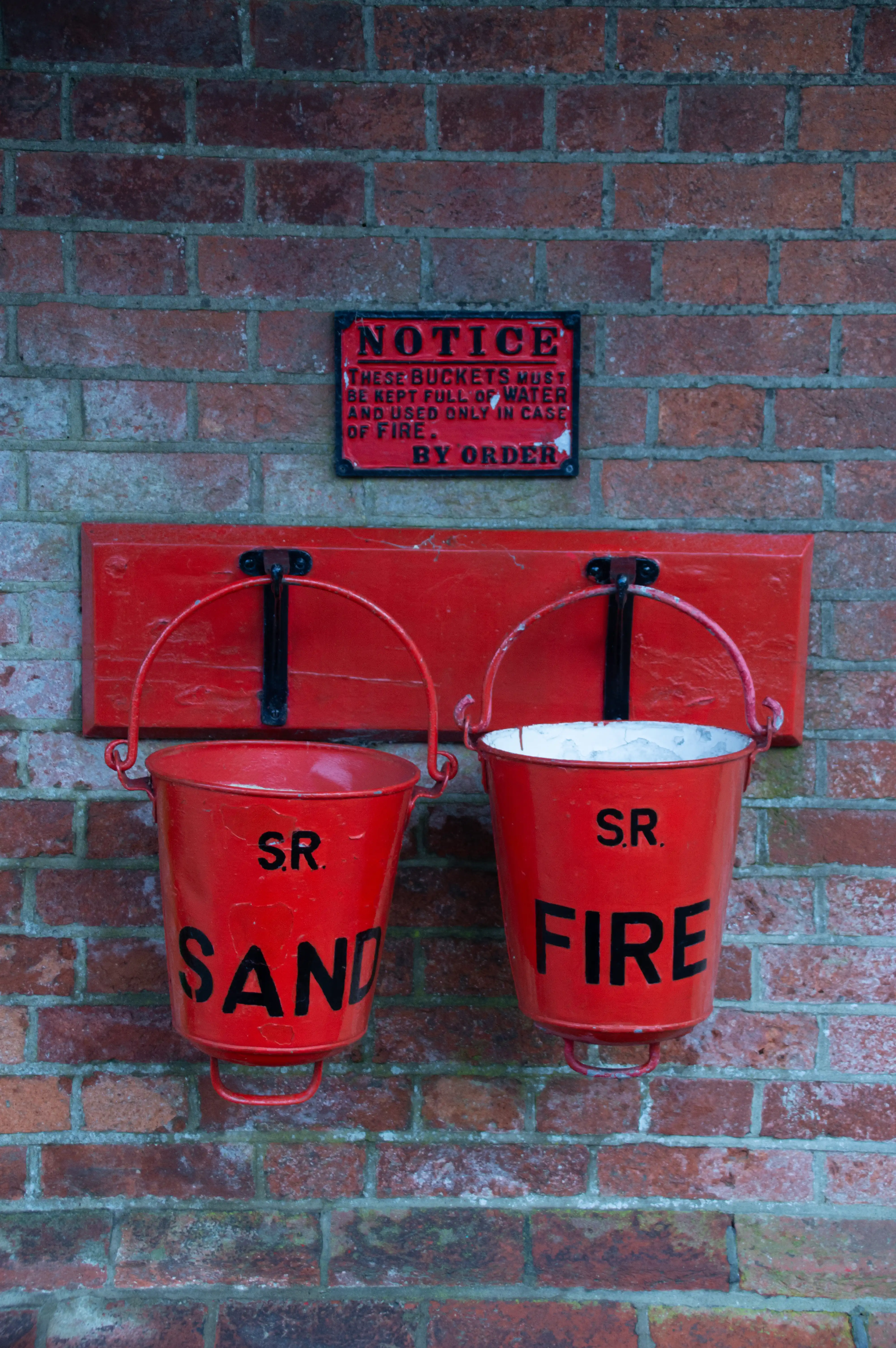 A close-up of two red metal fire buckets hanging on a brick wall, one labelled SAND and the other FIRE, with a notice above regarding their use.