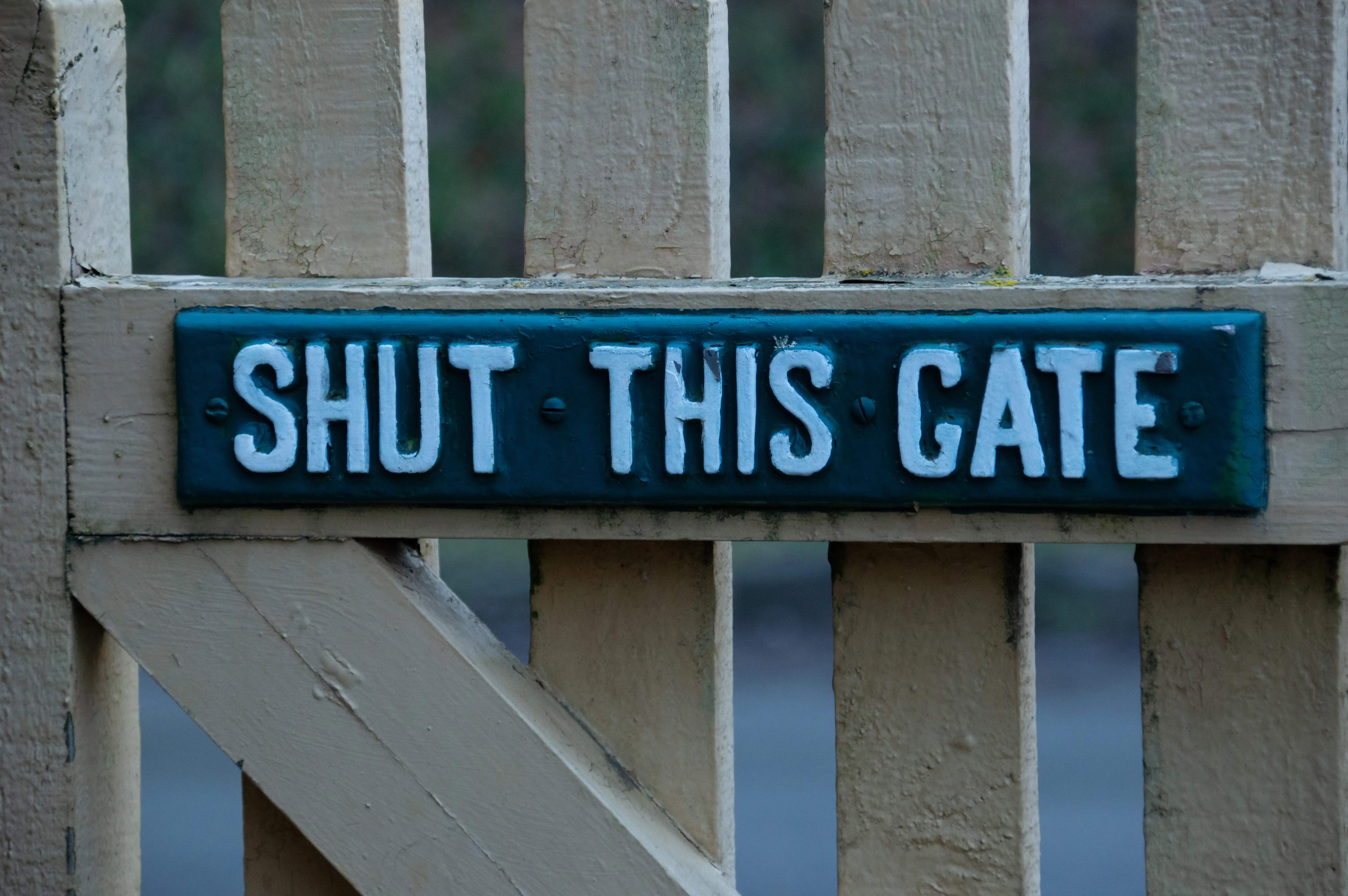 A close-up of a wooden gate with a turquoise plaque reading SHUT THIS GATE in raised silver letters.