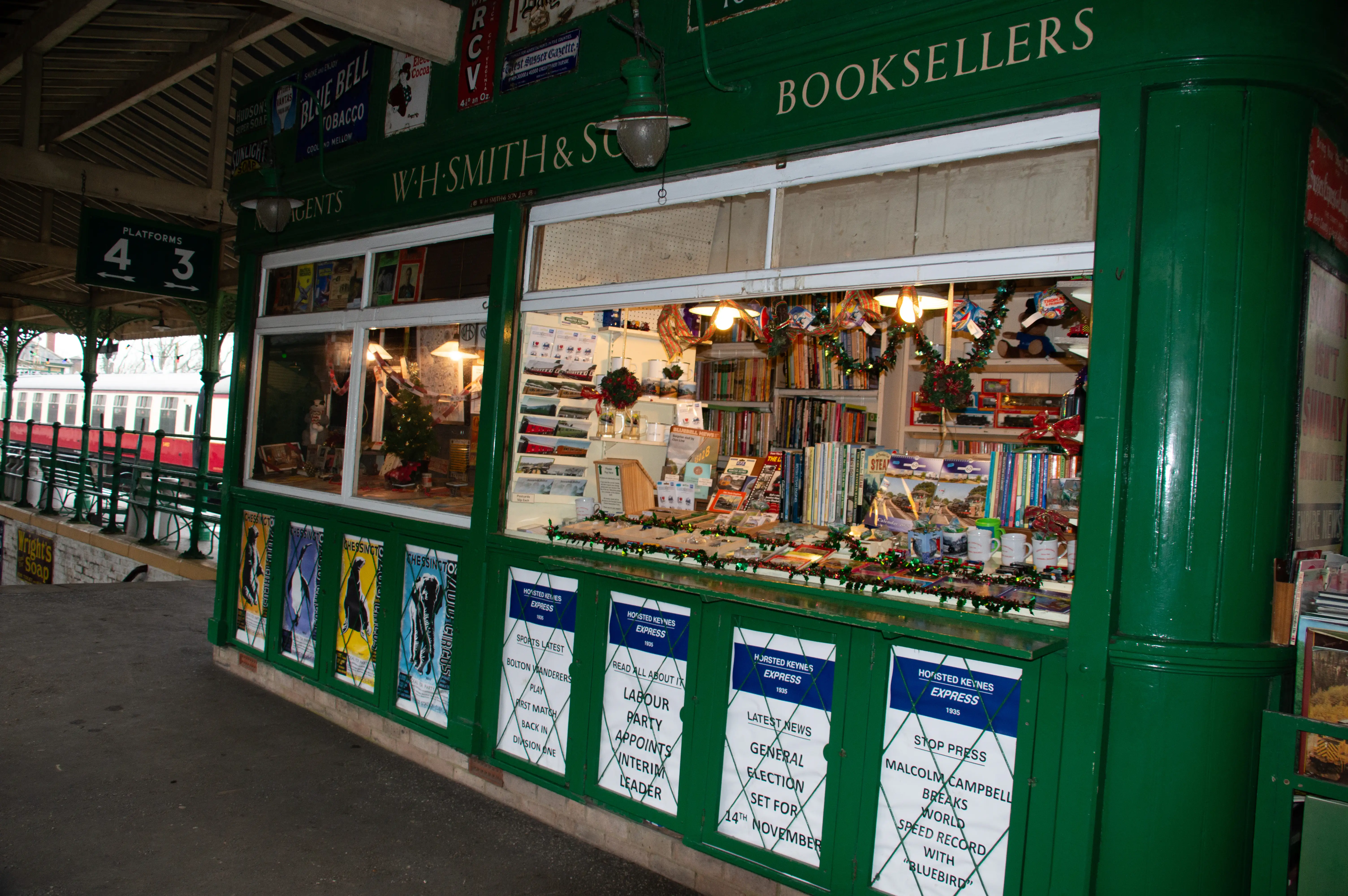 A green W.H. Smith bookstall at a train station, with its windows displaying books, Christmas decorations, and framed posters. Vintage advertisements are visible on the exterior walls, and a red and white train is parked on the platform to the left.
