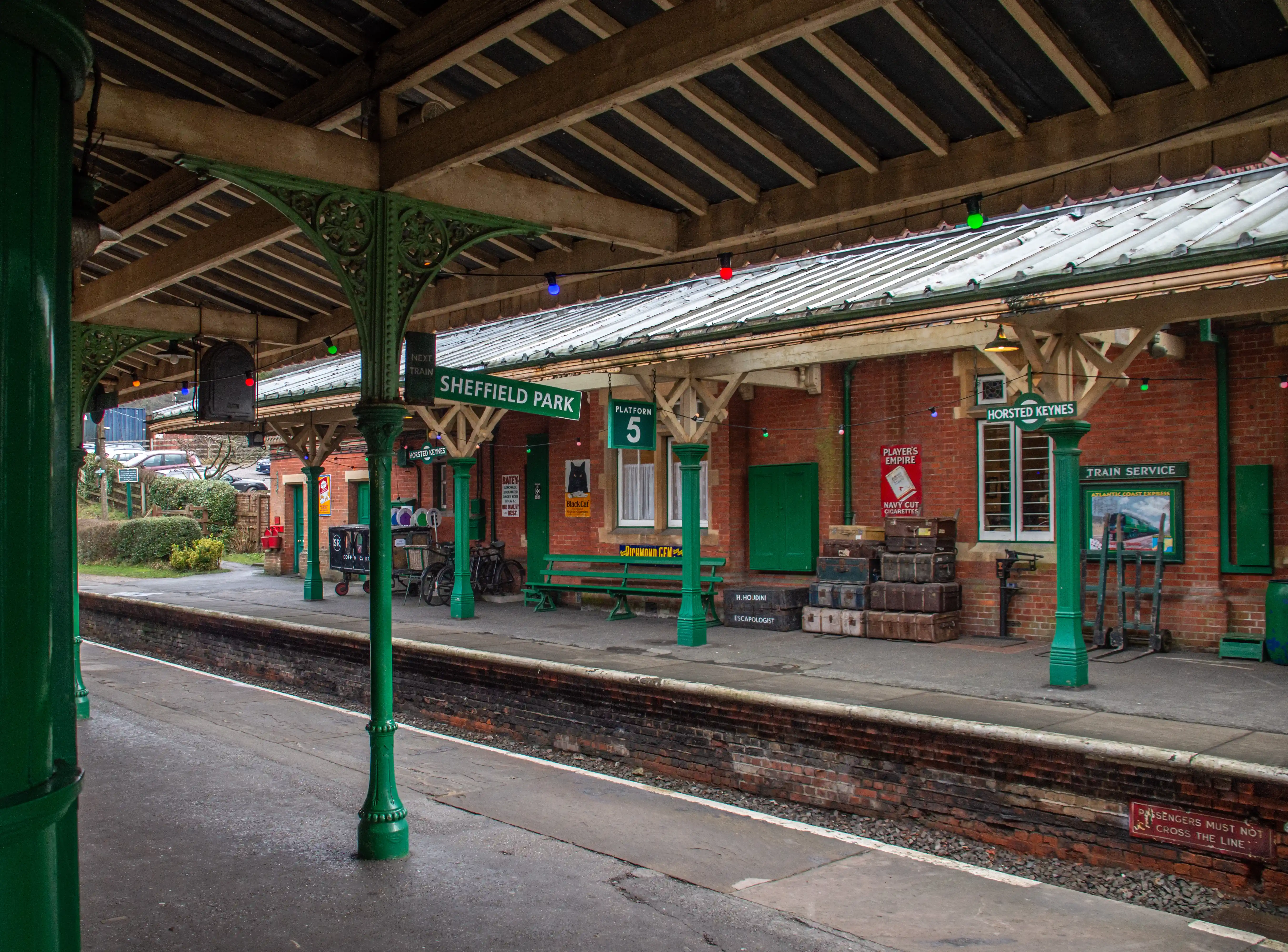A picturesque view of a historic train station platform with green painted columns and canopy. Vintage suitcases are stacked high on the platform, alongside a green wooden bench and bicycles. Signage indicates &laquo;Sheffield Park&raquo