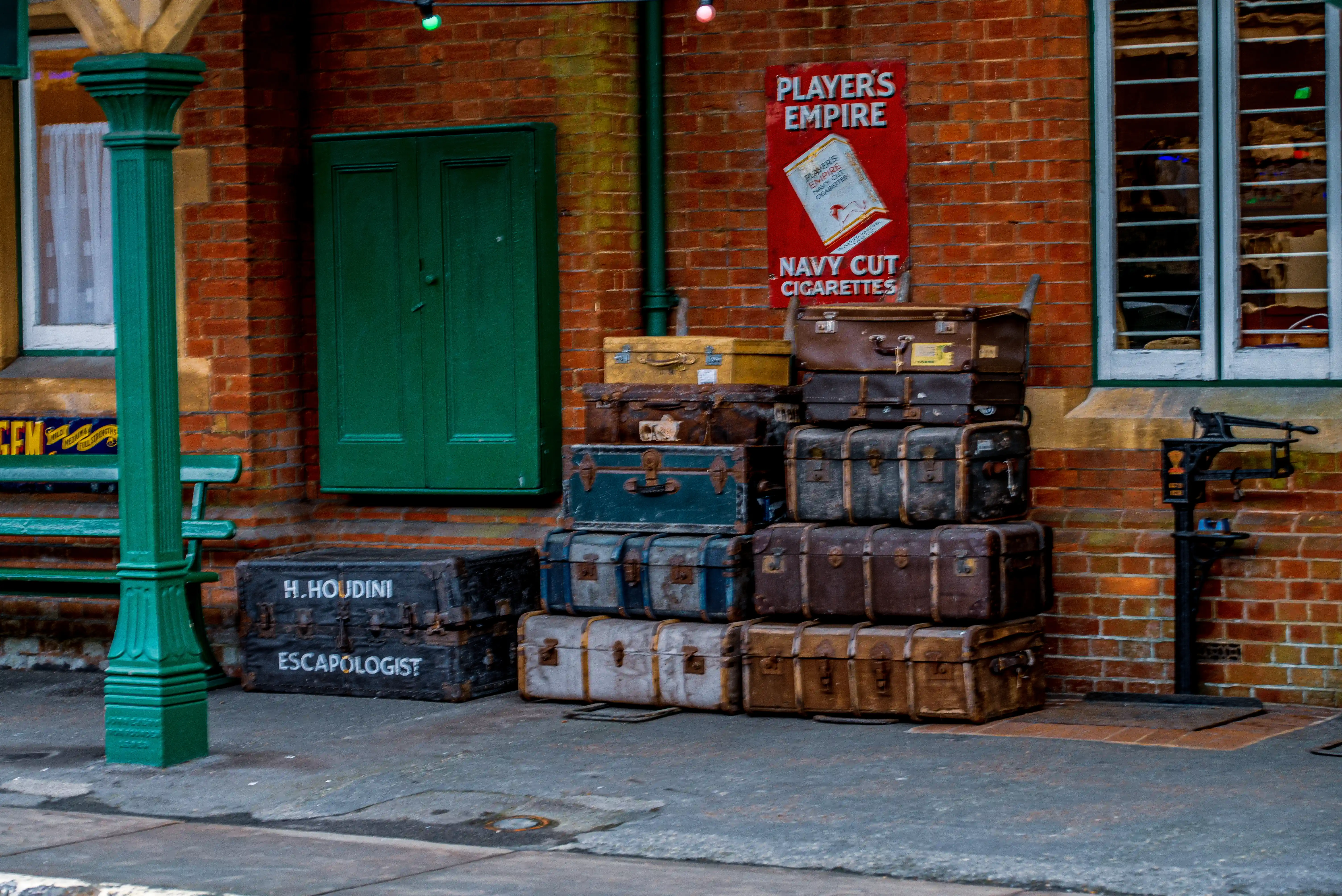 Stack of old suitcases, including one labelled 'H. Houdini Escapologist', with a vintage cigarette advert and a green door on a brick wall.