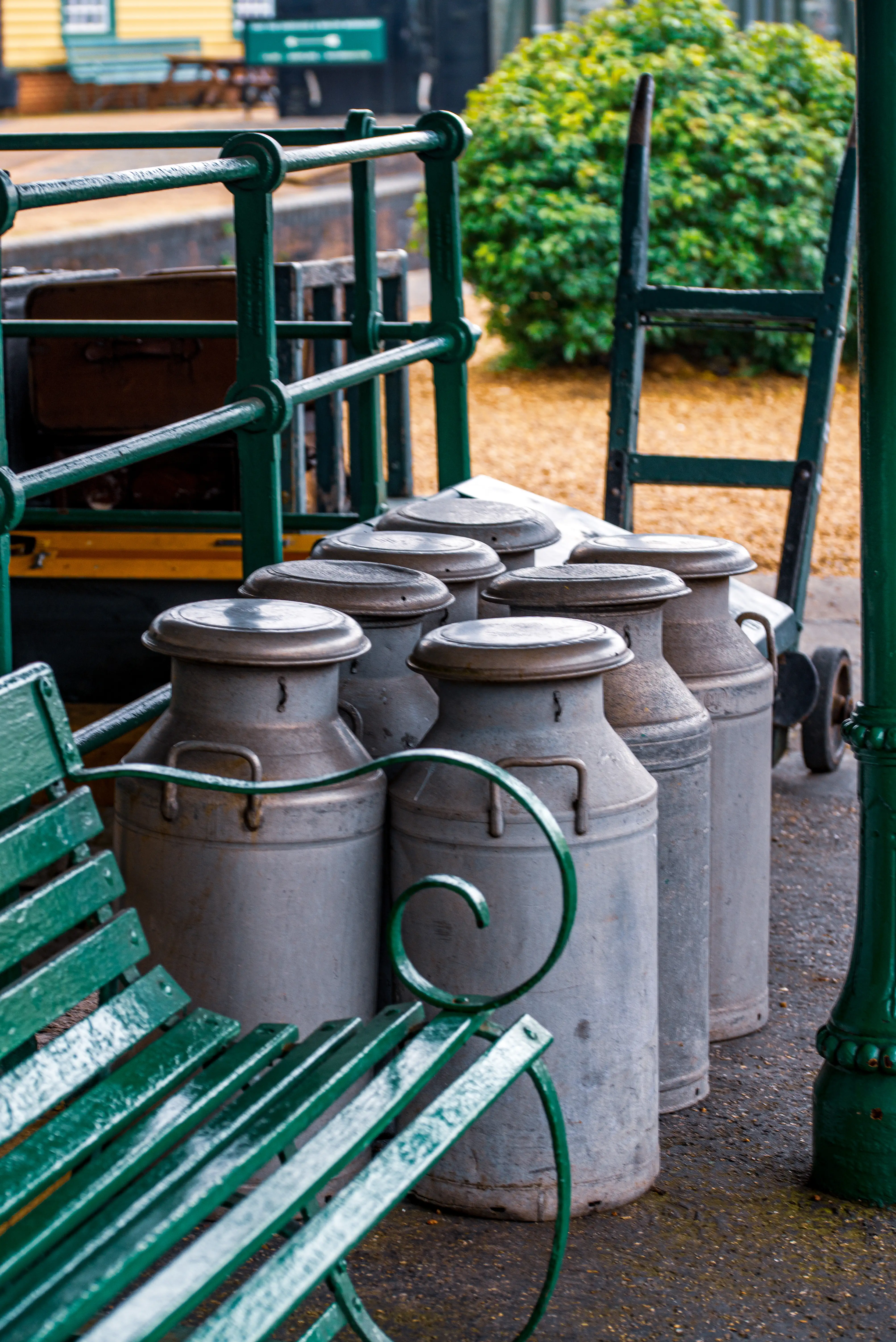 A group of large, metallic milk churns are lined up on a paved railway platform. A green painted wooden bench is in the foreground, and a ladder is visible behind the churns.