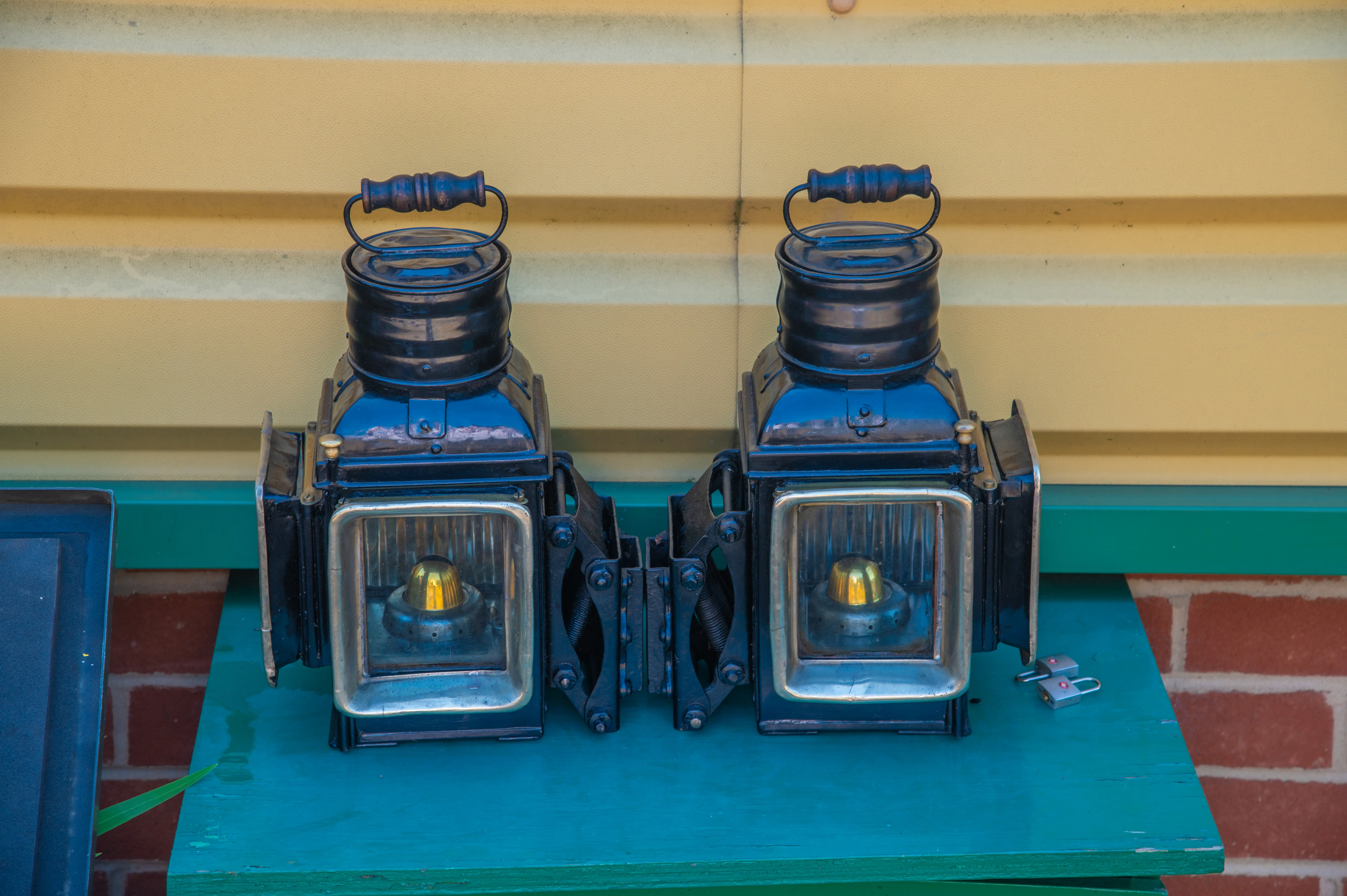 Two old-fashioned black train lanterns with yellow bulbs are displayed on a green shelf against a yellow corrugated wall.
