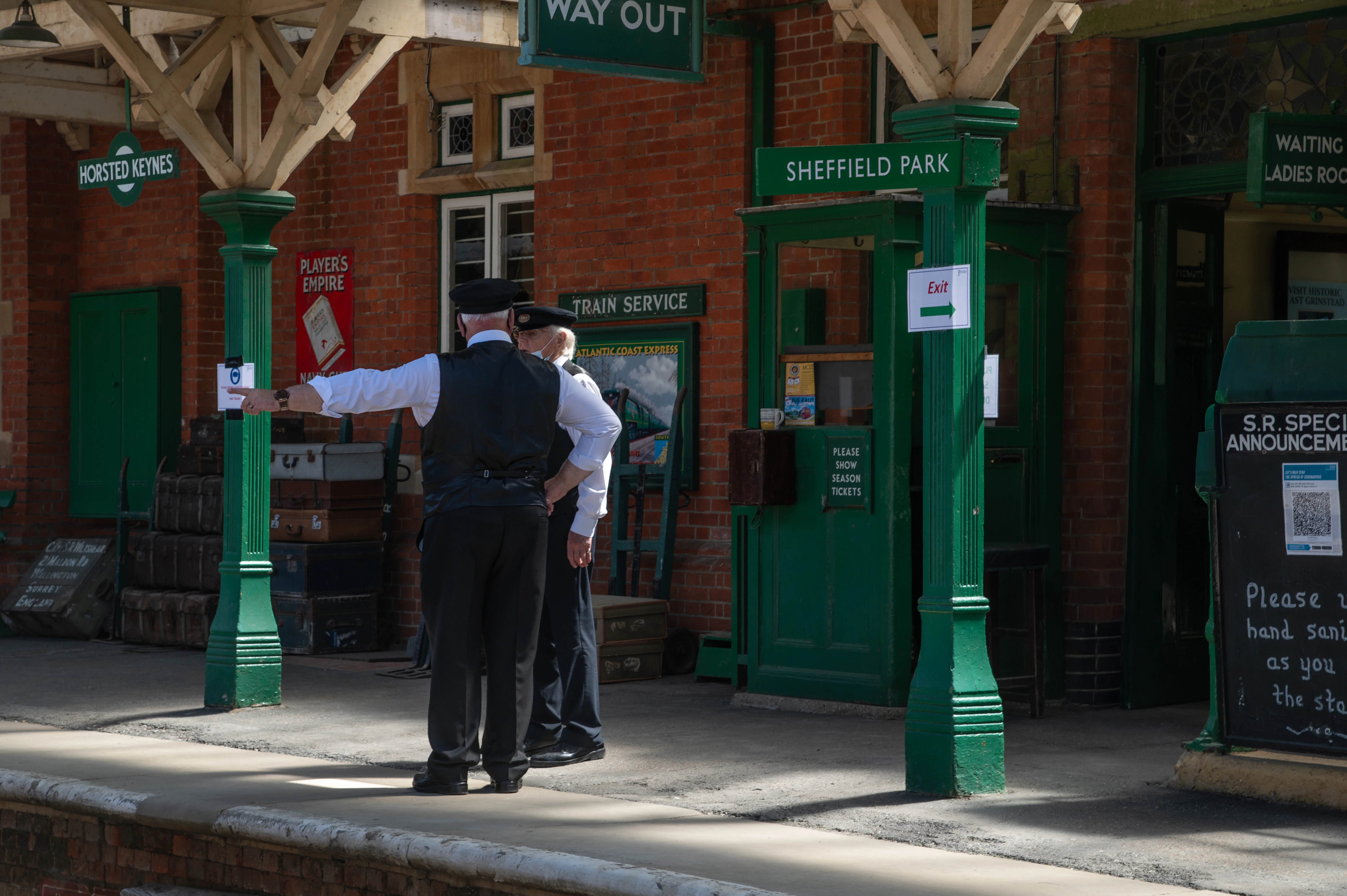 Two railway staff members in traditional uniforms standing on the platform at Horsted Keynes Station. One staff member points toward a sign, while the other observes. The station has green-painted wooden pillars with signs for 'WAY OUT' and 'SHEFFIELD PARK', a brick building, and vintage posters, including 'PLAYER'S EMPIRE' and 'ATLANTIC COAST EXPRESS'. A chalkboard sign reads: 'Please use hand sanitiser as you enter the station