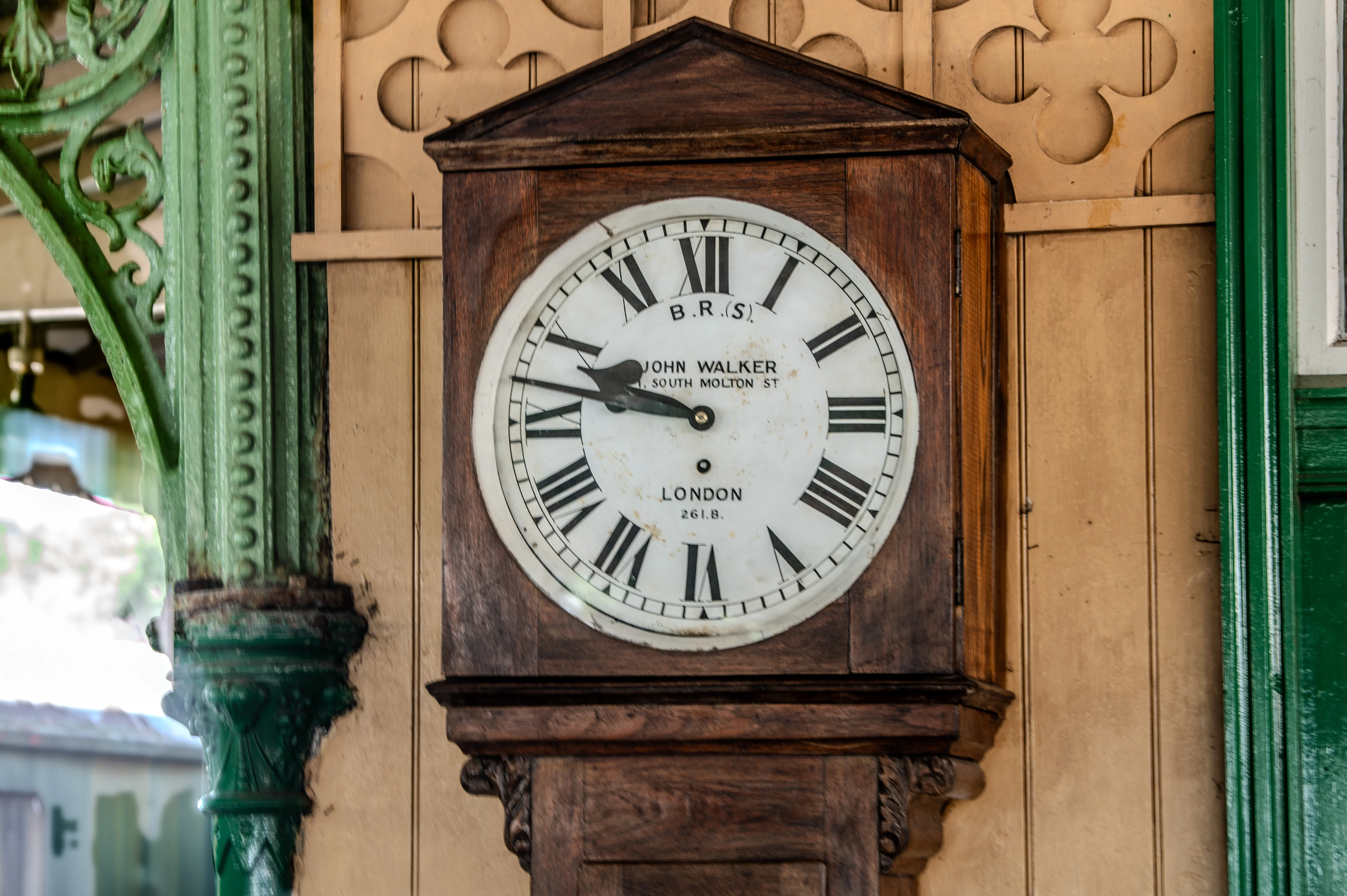 A detailed shot of an old wooden clock with a white face displaying Roman numerals and the maker's mark John Walker, London. The clock is set against a beige, wood-panelled background, with hints of green architectural elements on either side.