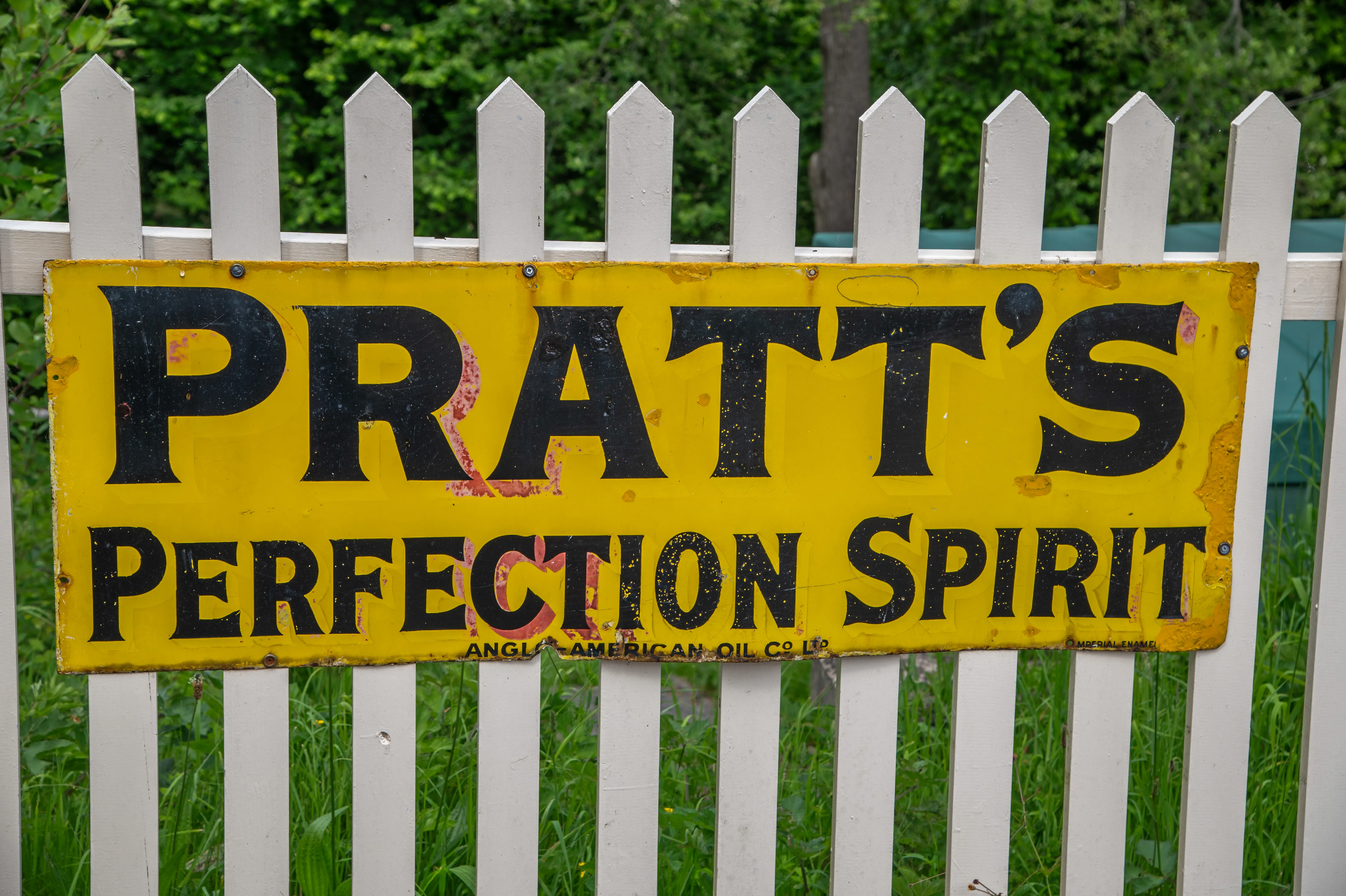A close-up of a vintage yellow enamel sign reading PRATT'S PERFECTION SPIRIT in black lettering, attached to a white picket fence with green foliage in the background.