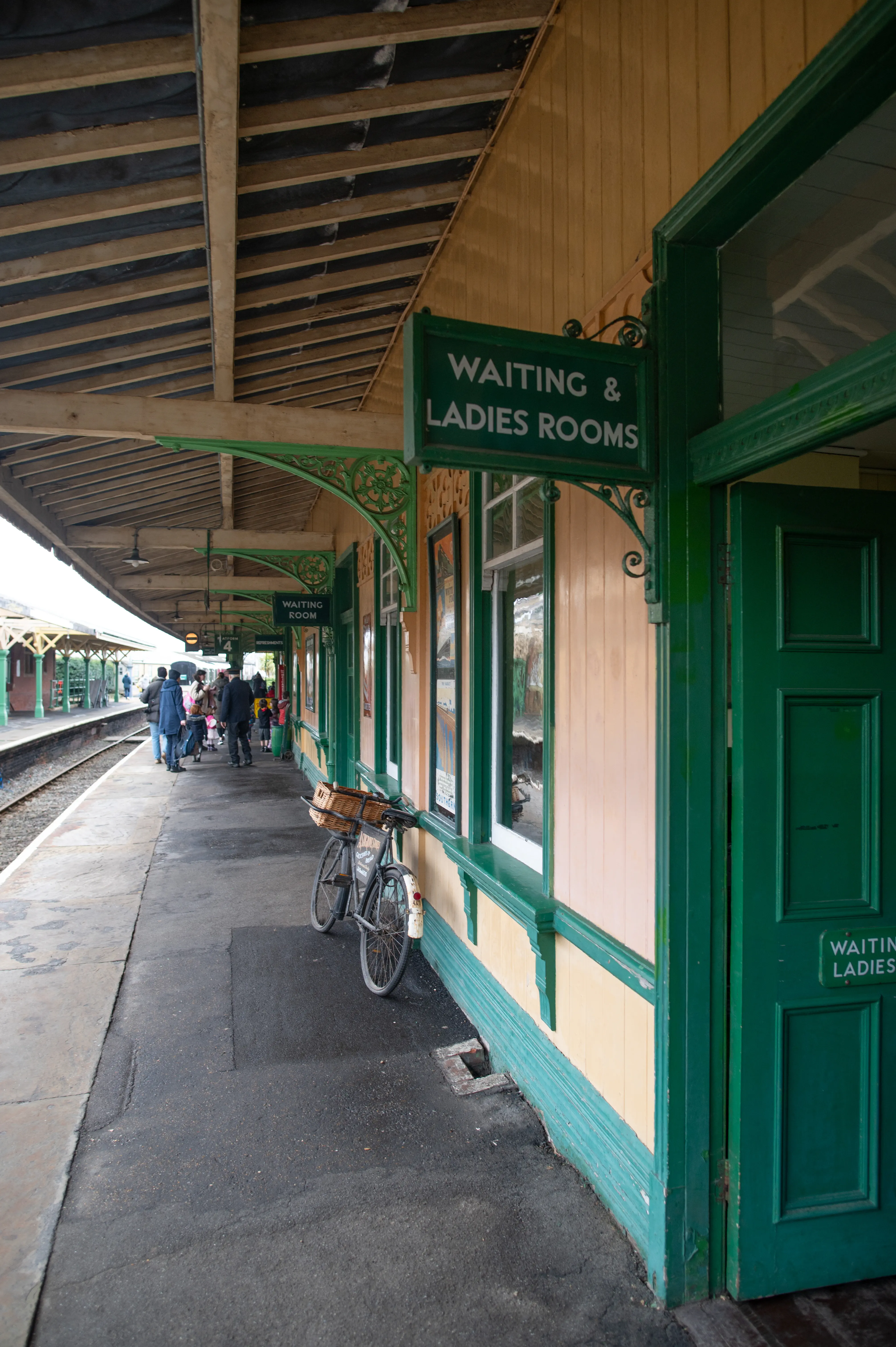 A side view of a traditional train station platform, with a wooden building painted in pale yellow and green, a prominent green sign for 'Waiting & Ladies Rooms', and a vintage bicycle with a wicker basket parked next to the building. Several people are visible in the background on the platform, and a train track runs alongside.